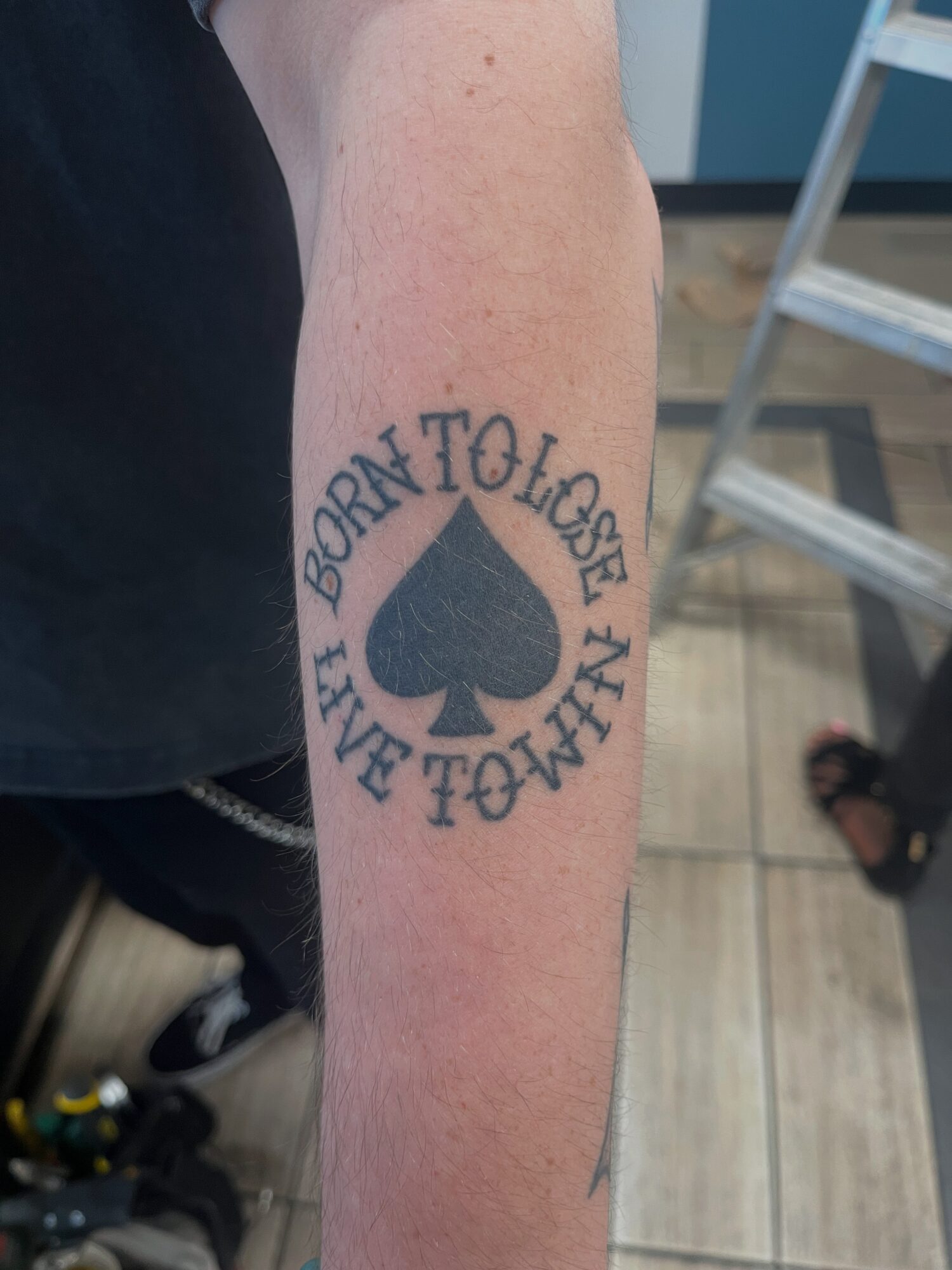 Tattoo of a spade symbol with the words 'BORN TO LOSE' and 'LIVE TO WIN' in a circular pattern on person's arm.