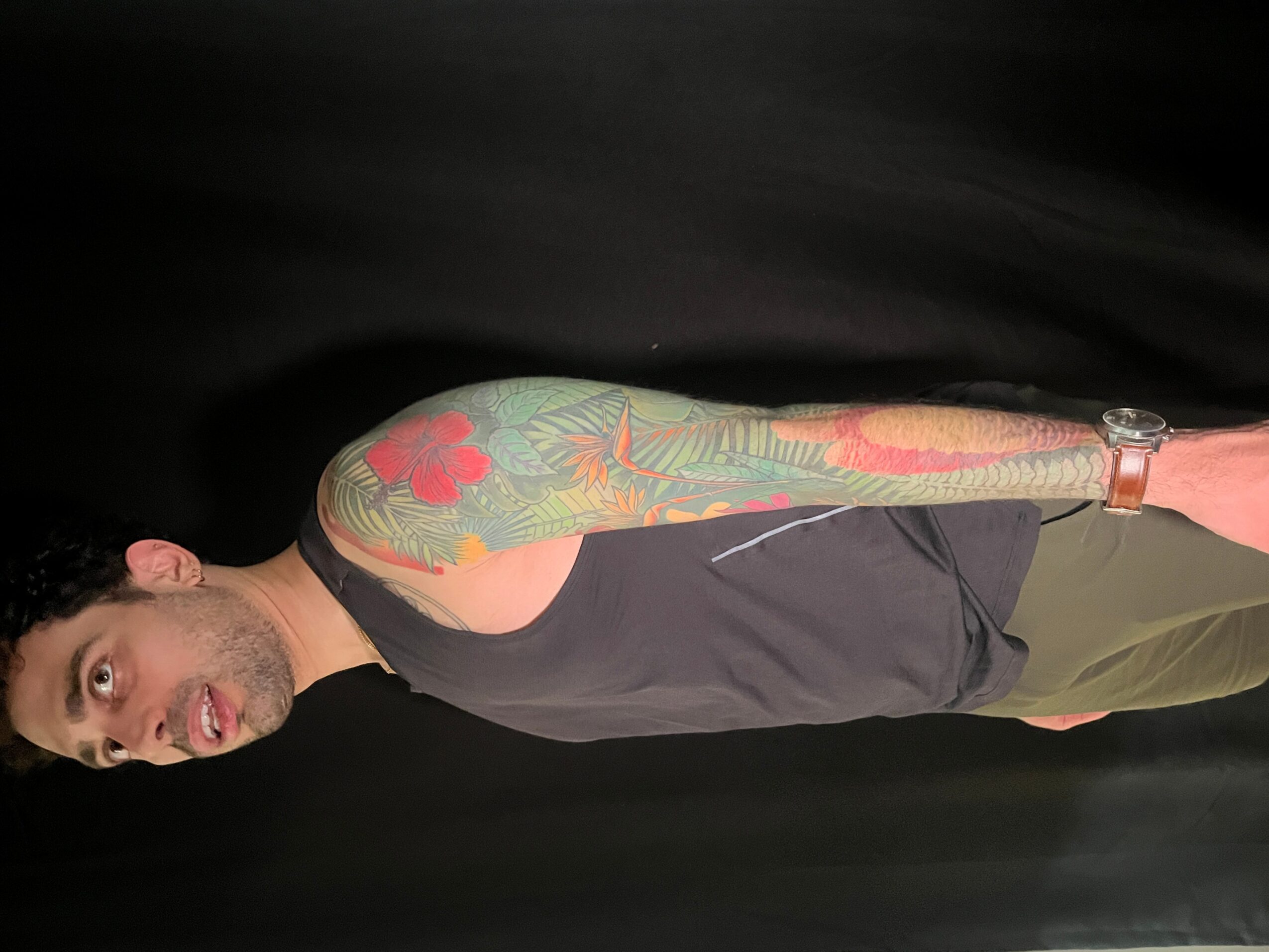 Man with colorful tattoo on arm, wearing black shirt and green pants, standing against black background.