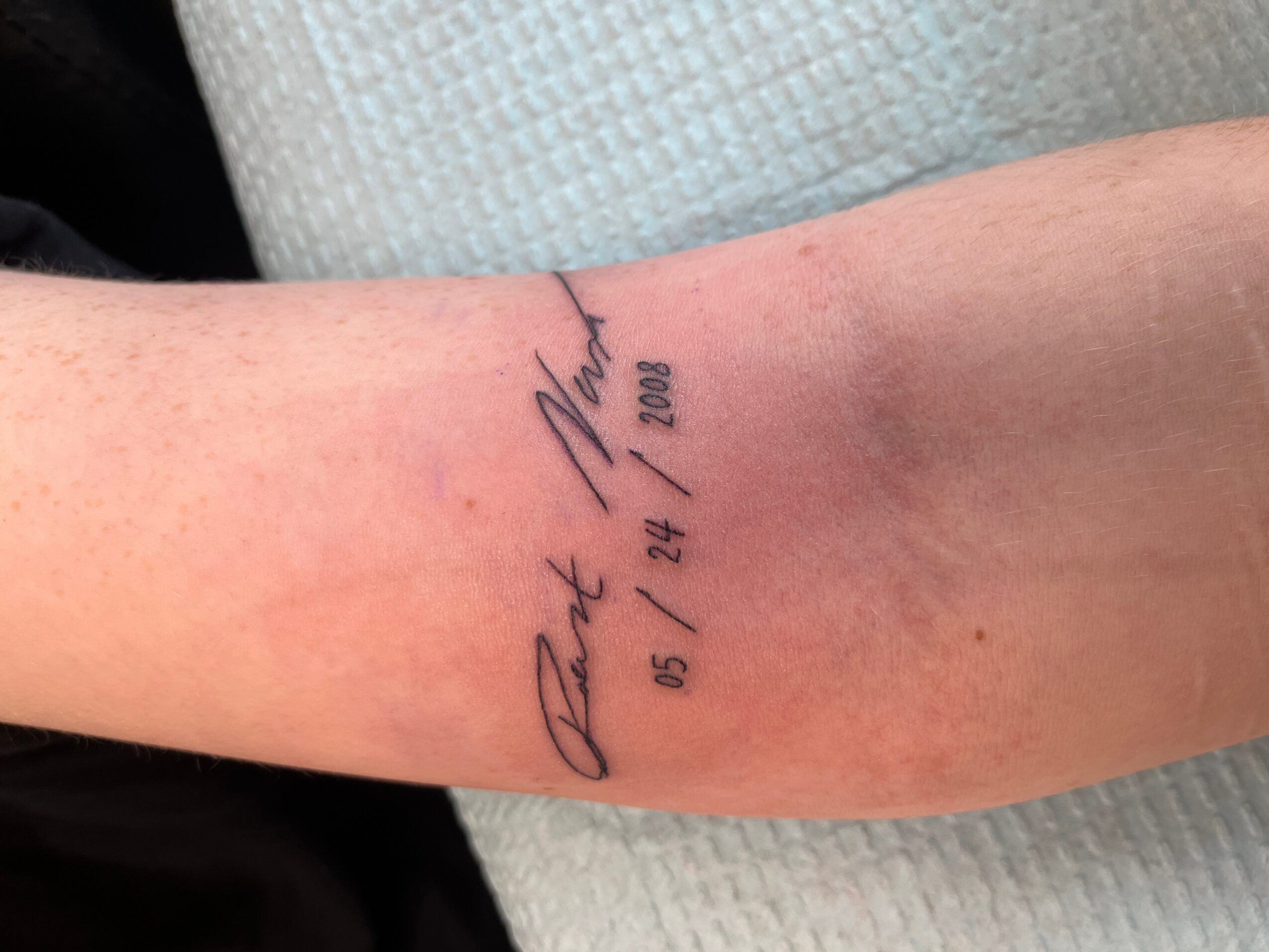 Tattoo on forearm with text and date, partially visible, against a light background.