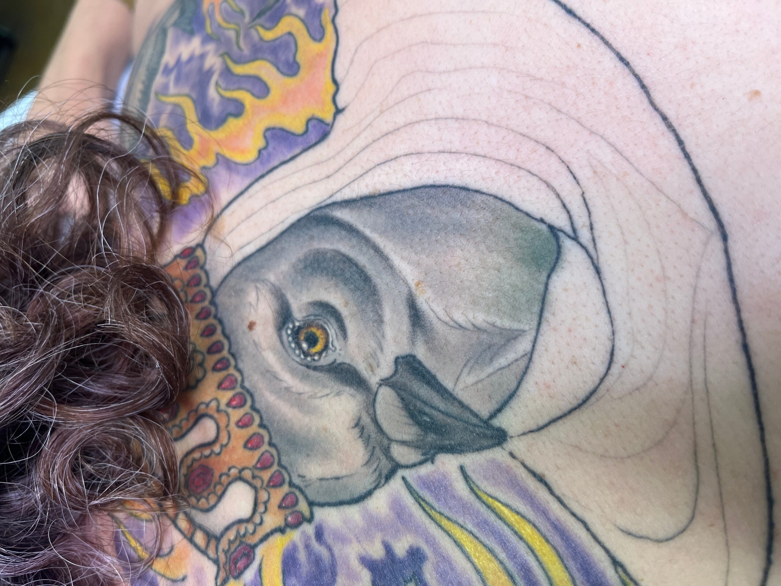 Person's back with a detailed, colorful tattoo of a face, surrounded by abstract patterns and designs.