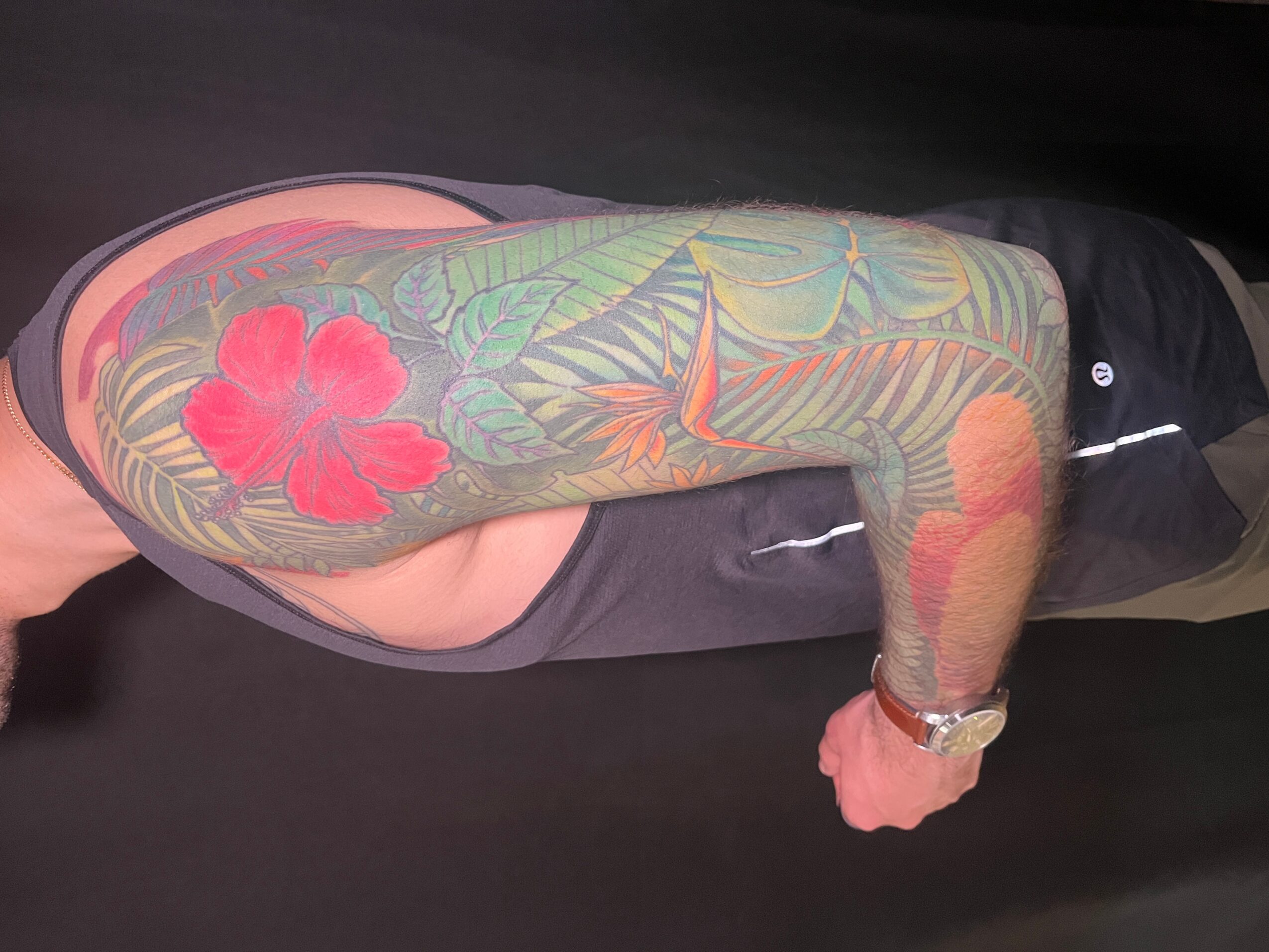 Person lying on side with colorful tattooed arm visible, wearing a watch, dark clothing, and a black background.