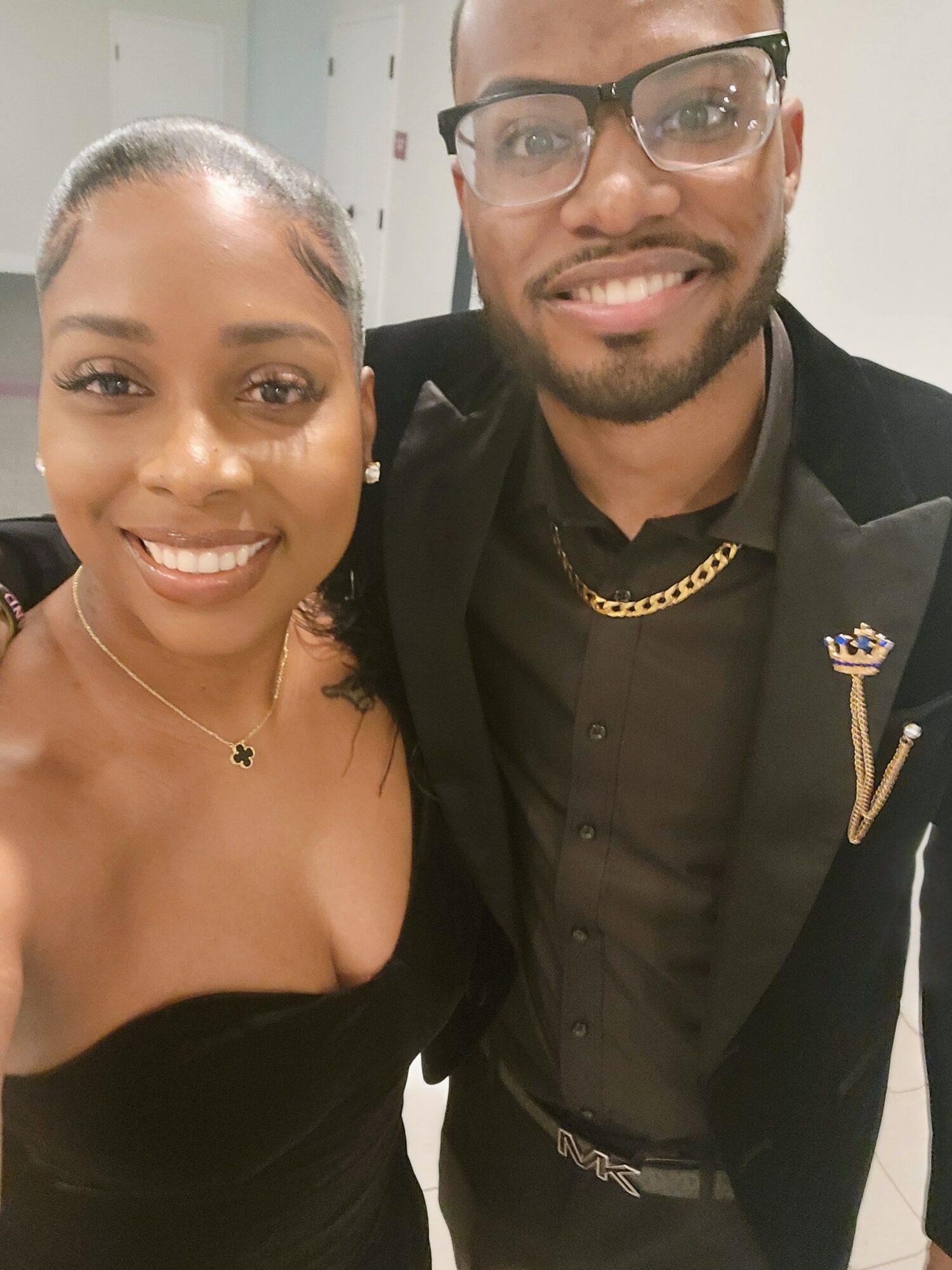 Two smiling people, one woman with a necklace and tattoo, and one man with glasses and a chain, pose together indoors.