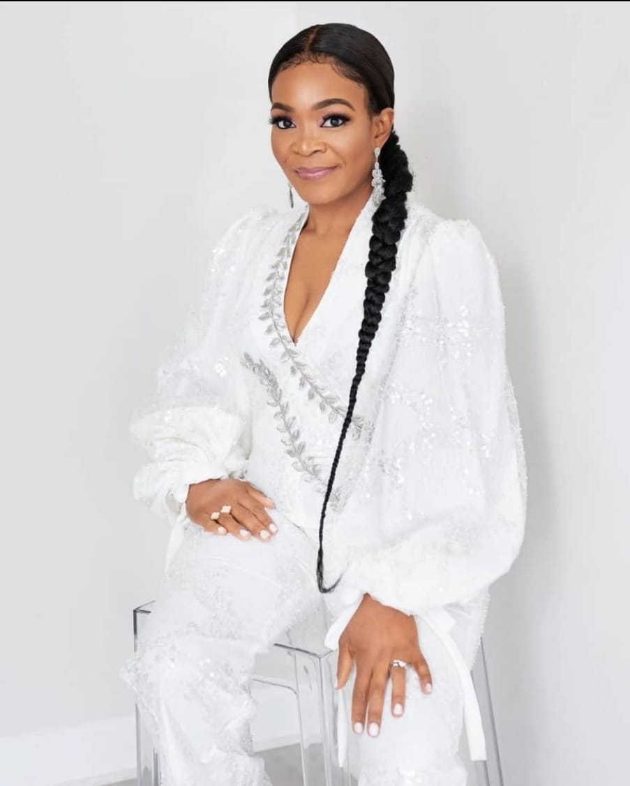 Woman with long braided hair wearing a white textured dress, standing against a plain background.