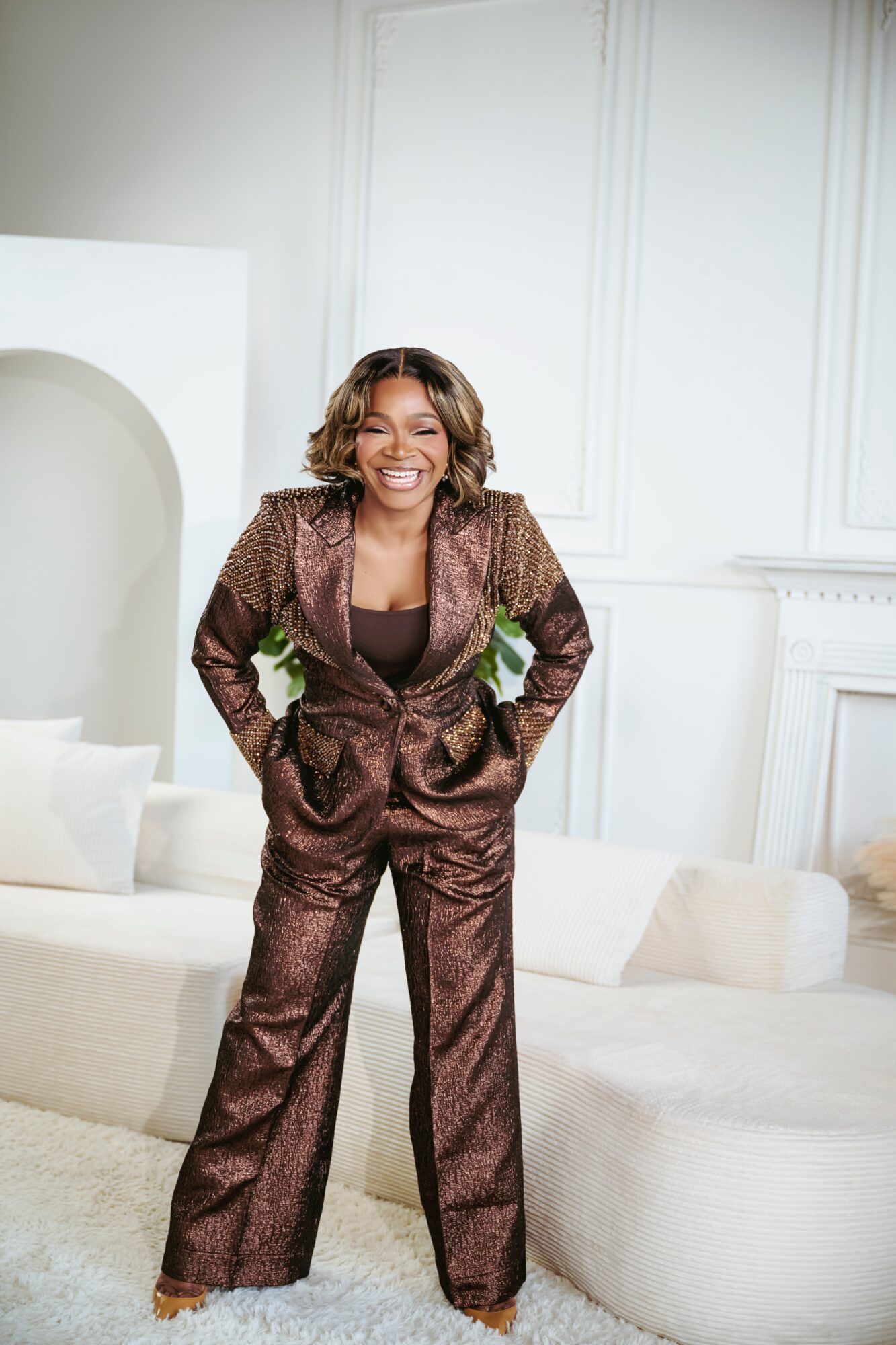 Woman with curly hair smiling, hands in pockets, standing in a bright room with white walls and furniture.