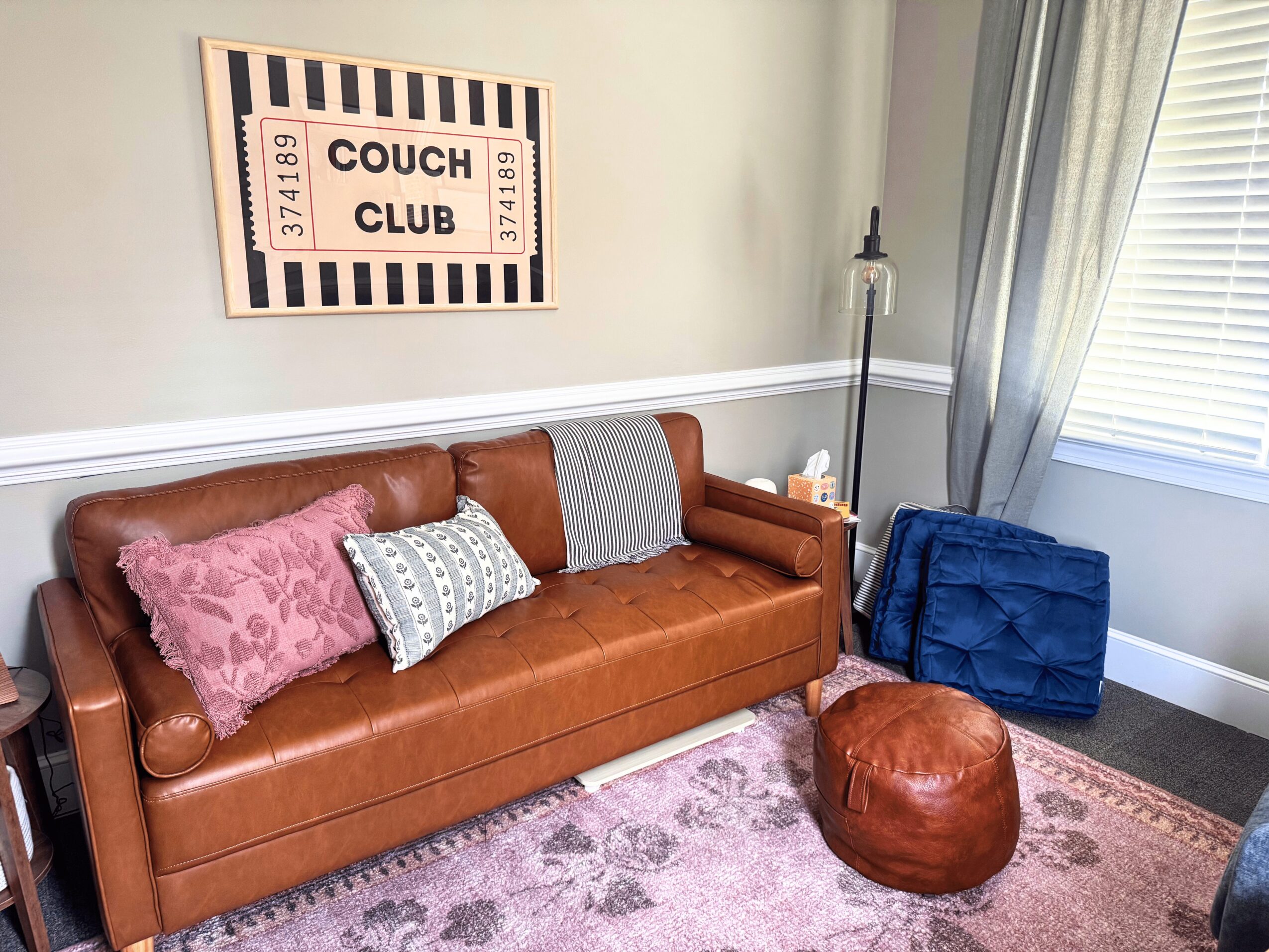Living room with a brown leather sofa, pink and patterned pillows, a leather ottoman, a floor lamp, and a window with curtains.