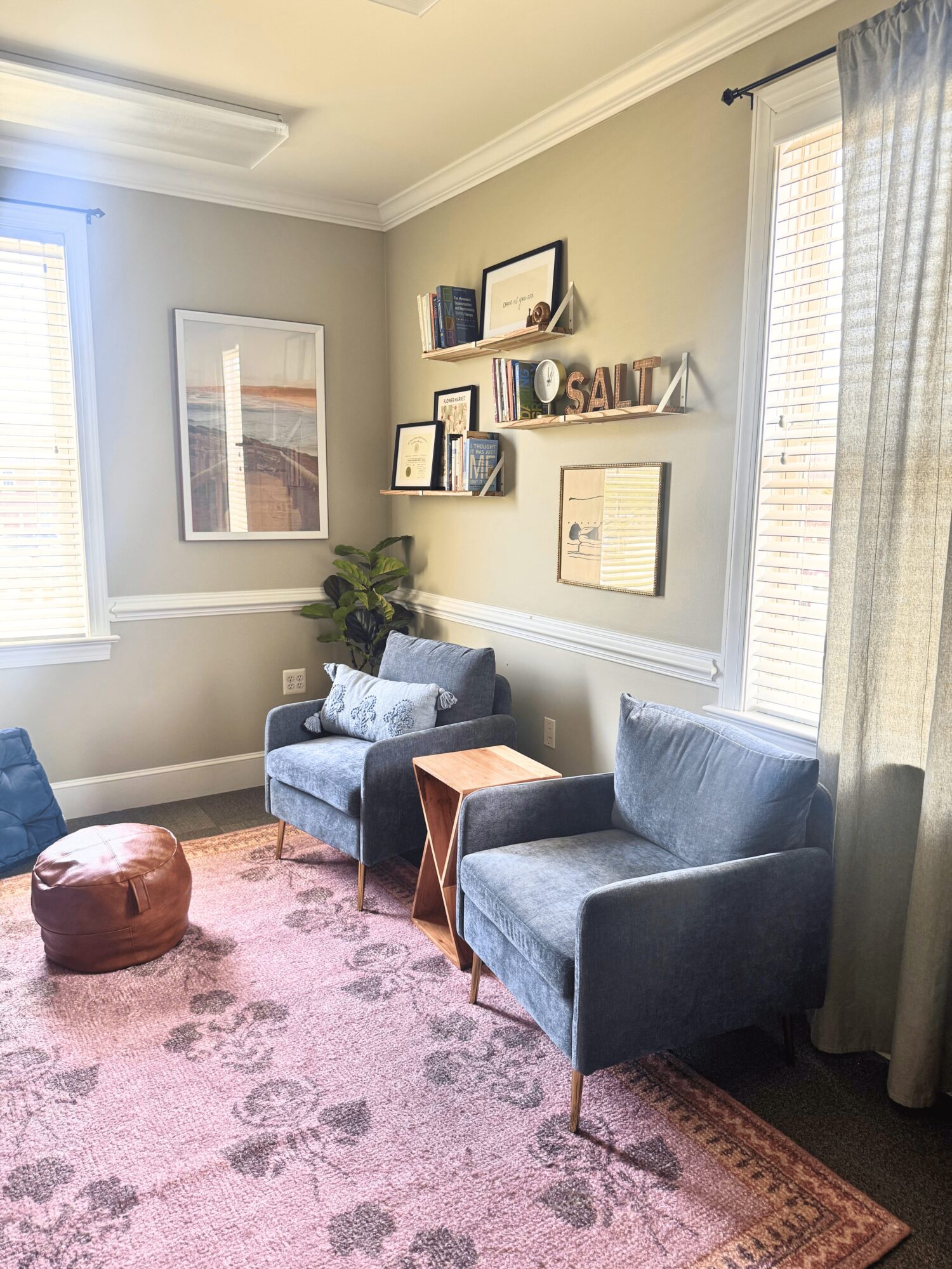 Corner of a room with two blue armchairs, a small wooden table, a pink pouf, and wall shelves with books and decor, sunlight streaming through windows.