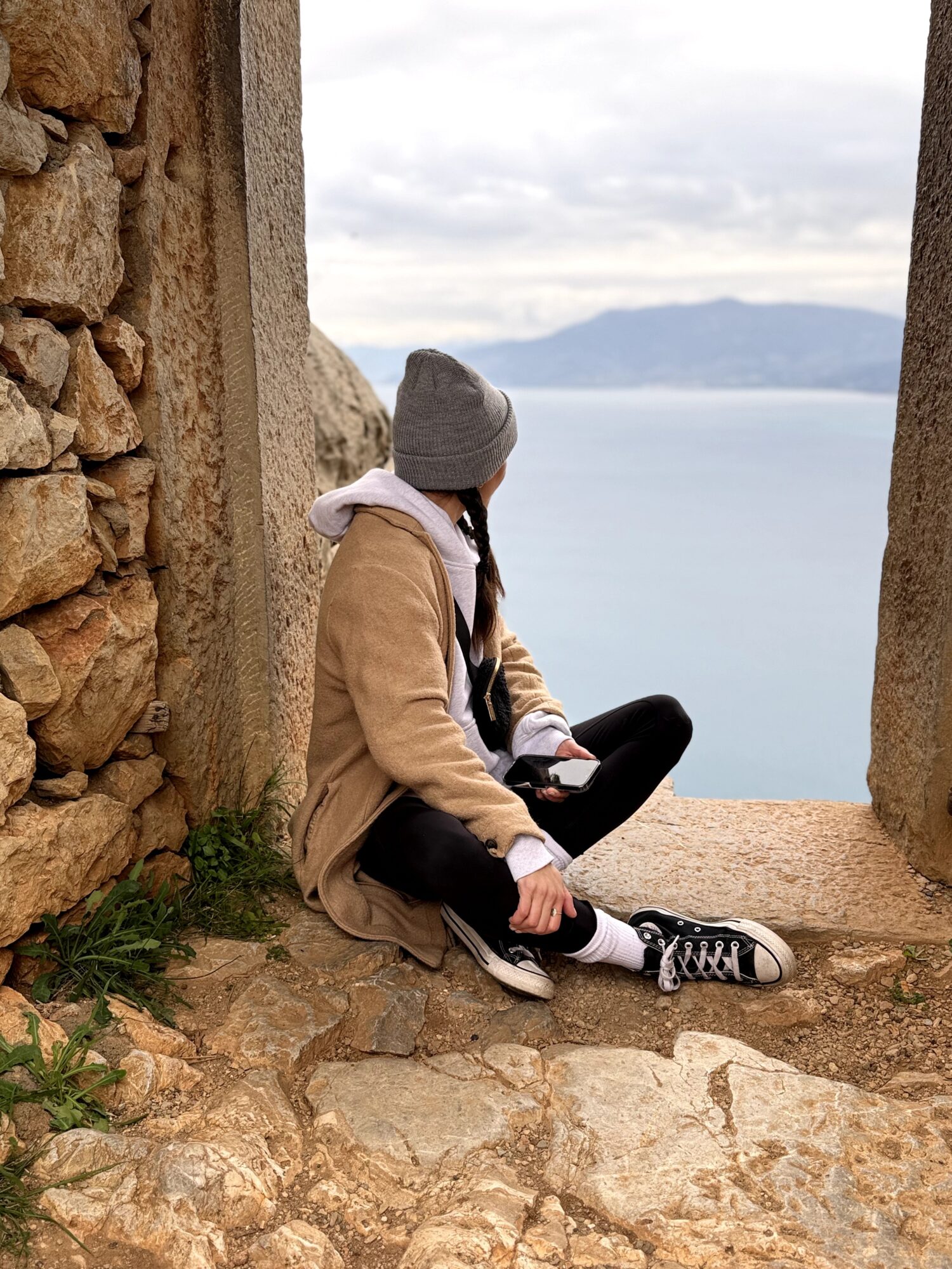 Person sitting on stone ground between stone and concrete walls, overlooking water and distant mountains, wearing a gray beanie and beige coat.