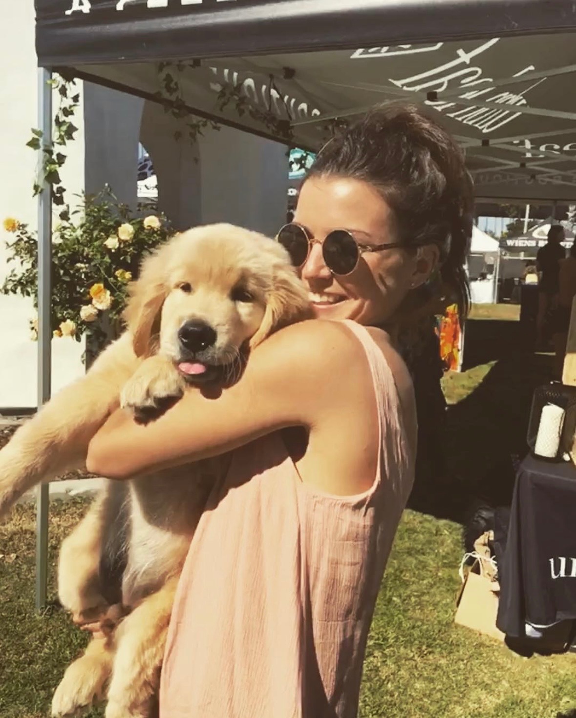 Woman with sunglasses holding a golden retriever puppy outdoors, smiling, under a canopy with flowers and tents nearby.