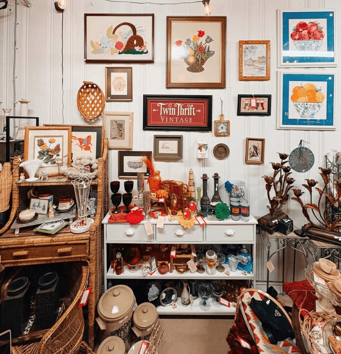 Wall decorated with framed artwork, shelves, and vintage items; white shelf with collectibles and bottles; various objects and plants on tables.