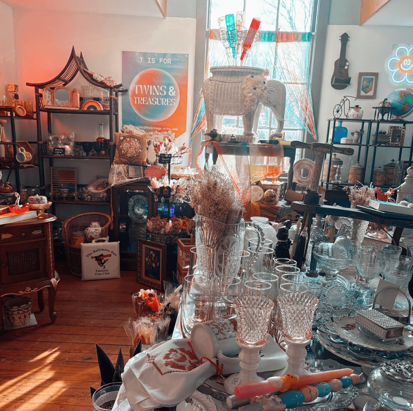 Room filled with various decorative items, glassware, and furniture, with sunlight streaming through a window, creating a cozy atmosphere.