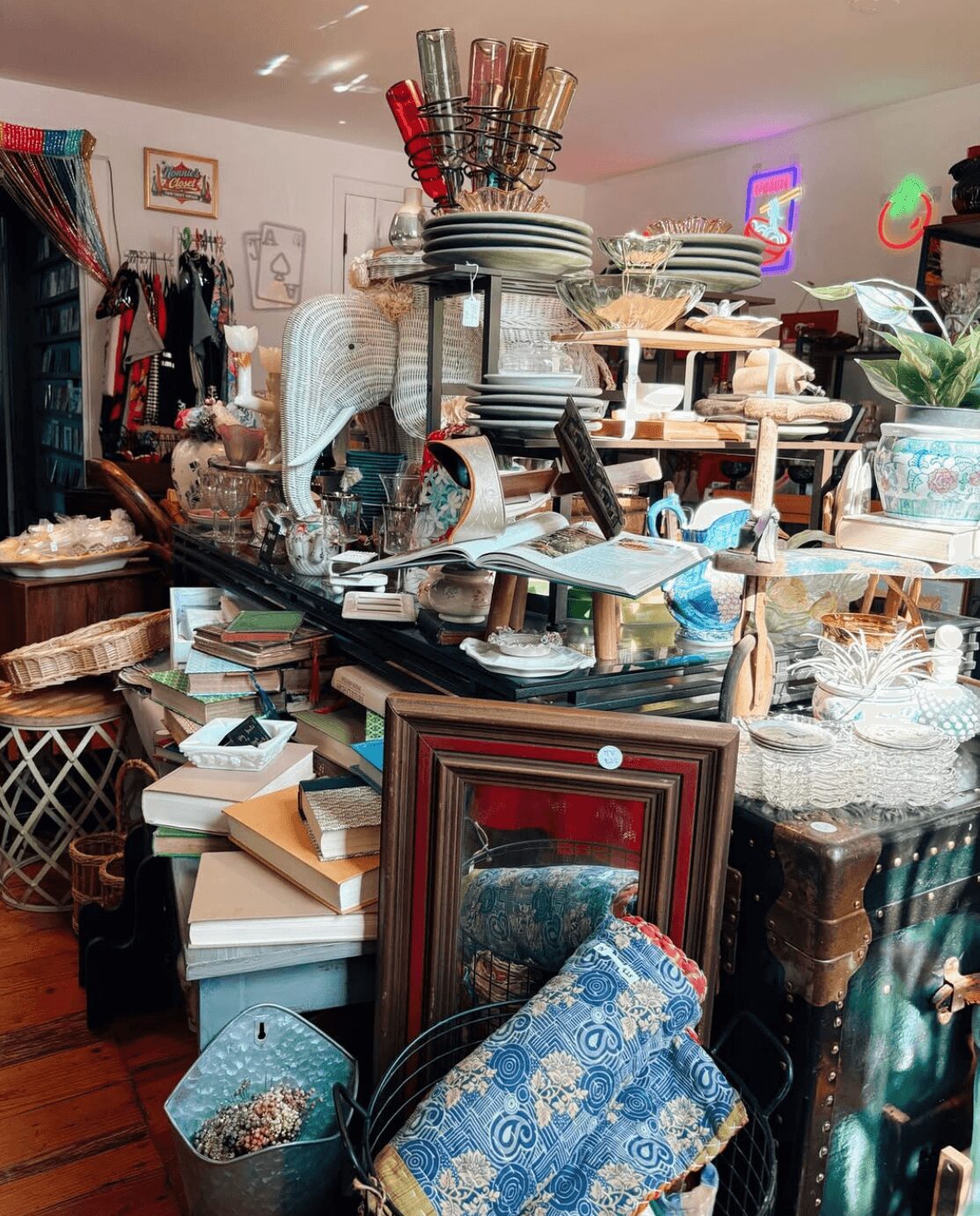 A cluttered room with shelves and tables filled with books, picture frames, and decorative items, including glassware and small furniture pieces.