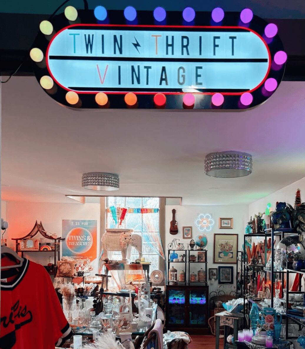 Interior of a vintage store with shelves, tables, and colorful decorations, under a neon sign reading 'Twin Thrift Vintage'.