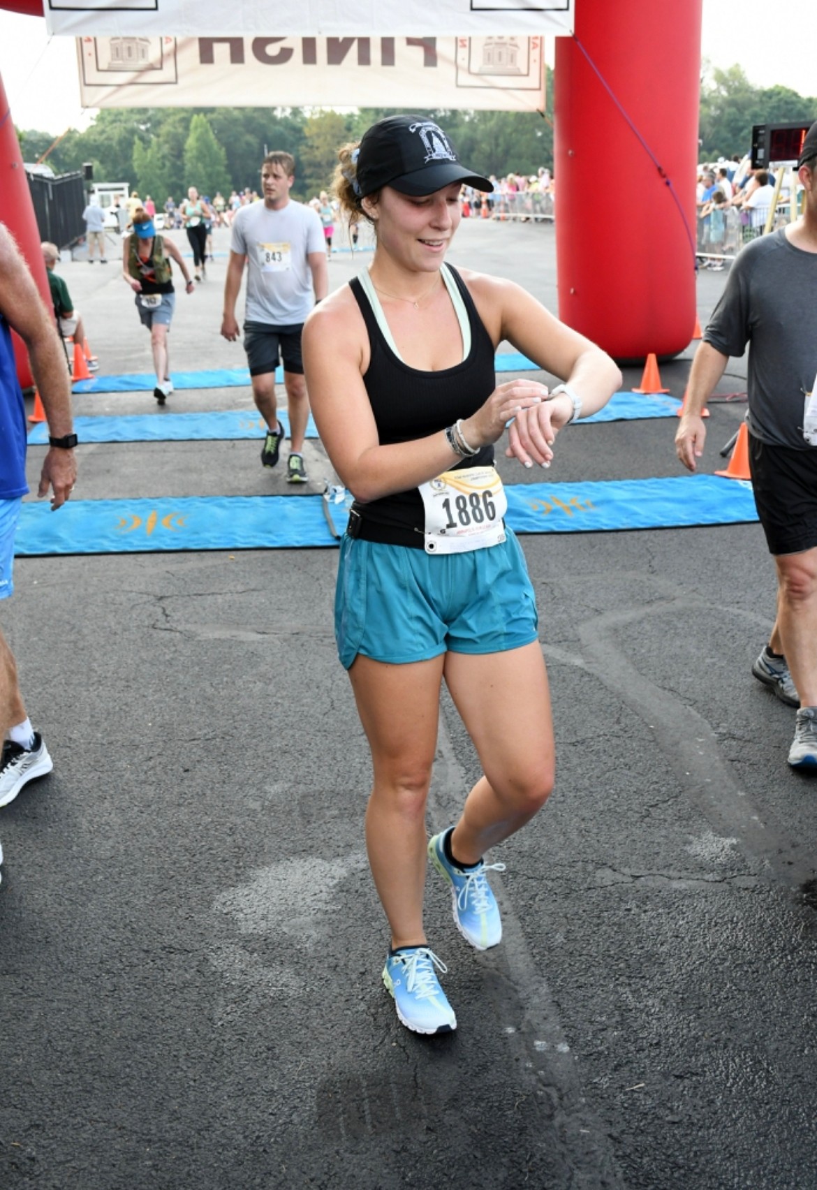Woman in athletic clothing checking her watch at a race finish line, with other runners and a red arch in background.