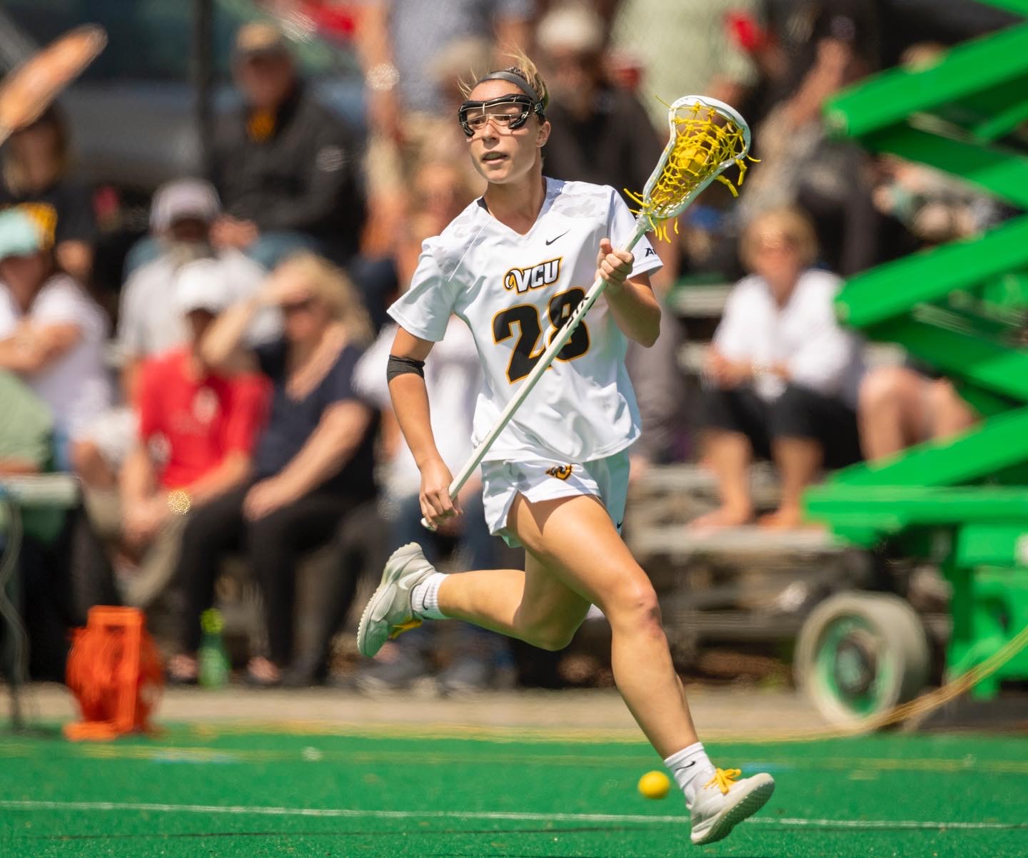 Female lacrosse player running on field with crowd in background, holding a yellow lacrosse stick, wearing a white uniform with number 22.