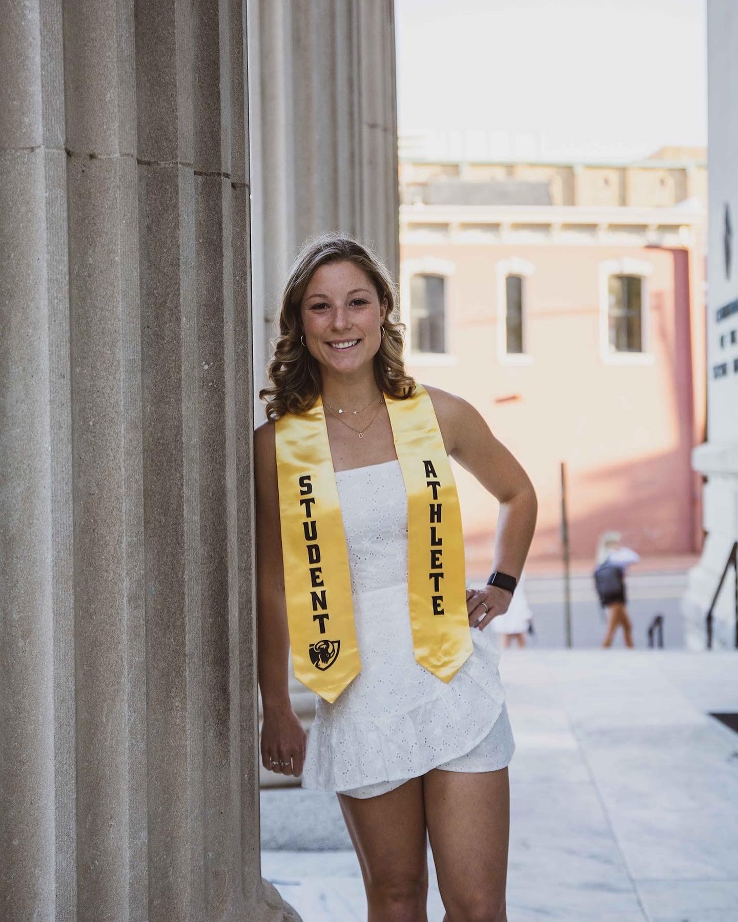 Young woman smiling, wearing a yellow sash with 'STUDENT ATHLETE', standing outdoors near a stone column.