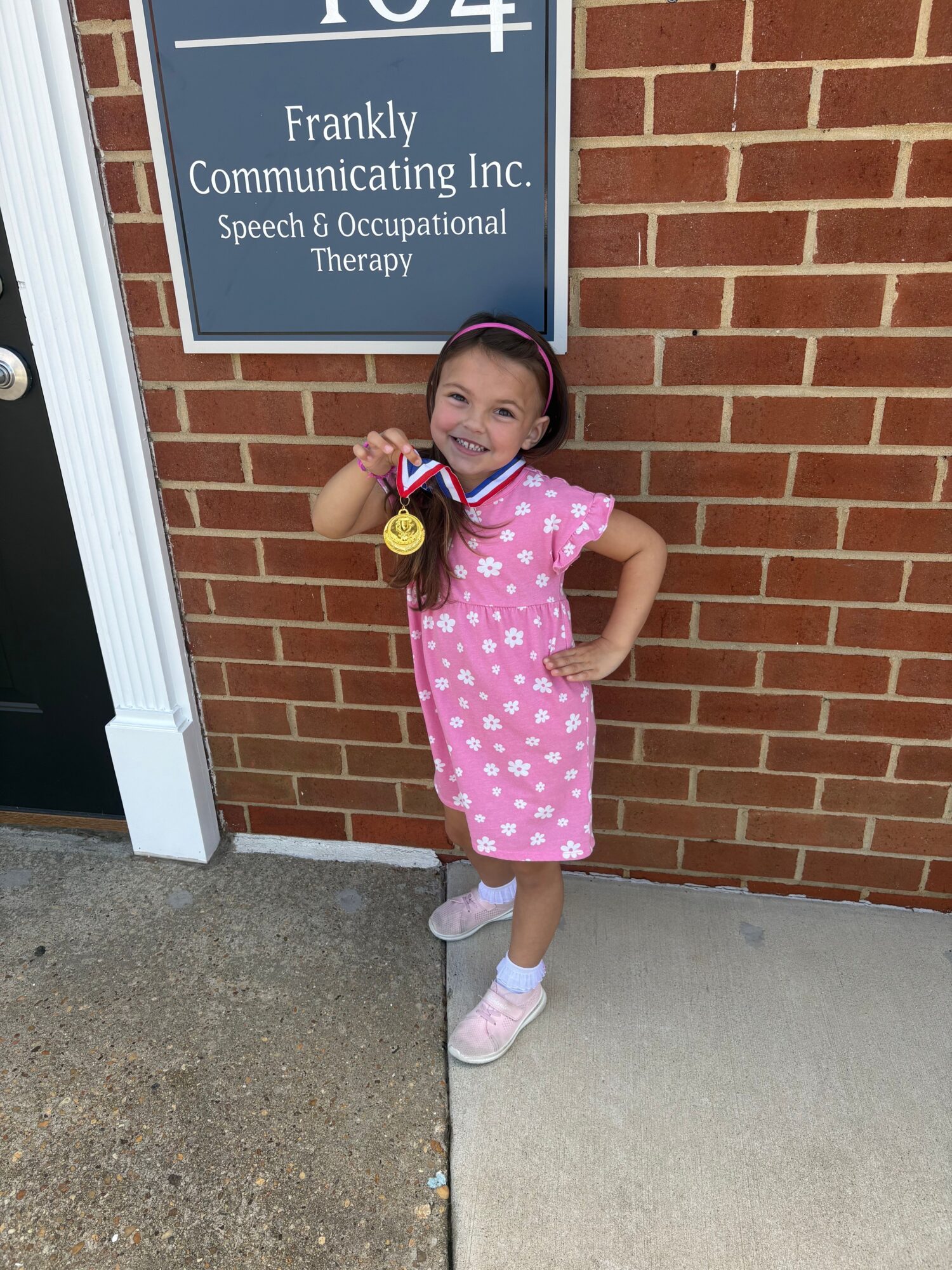 Young girl in pink polka dot dress holding a toy, standing against a brick wall outside a building.