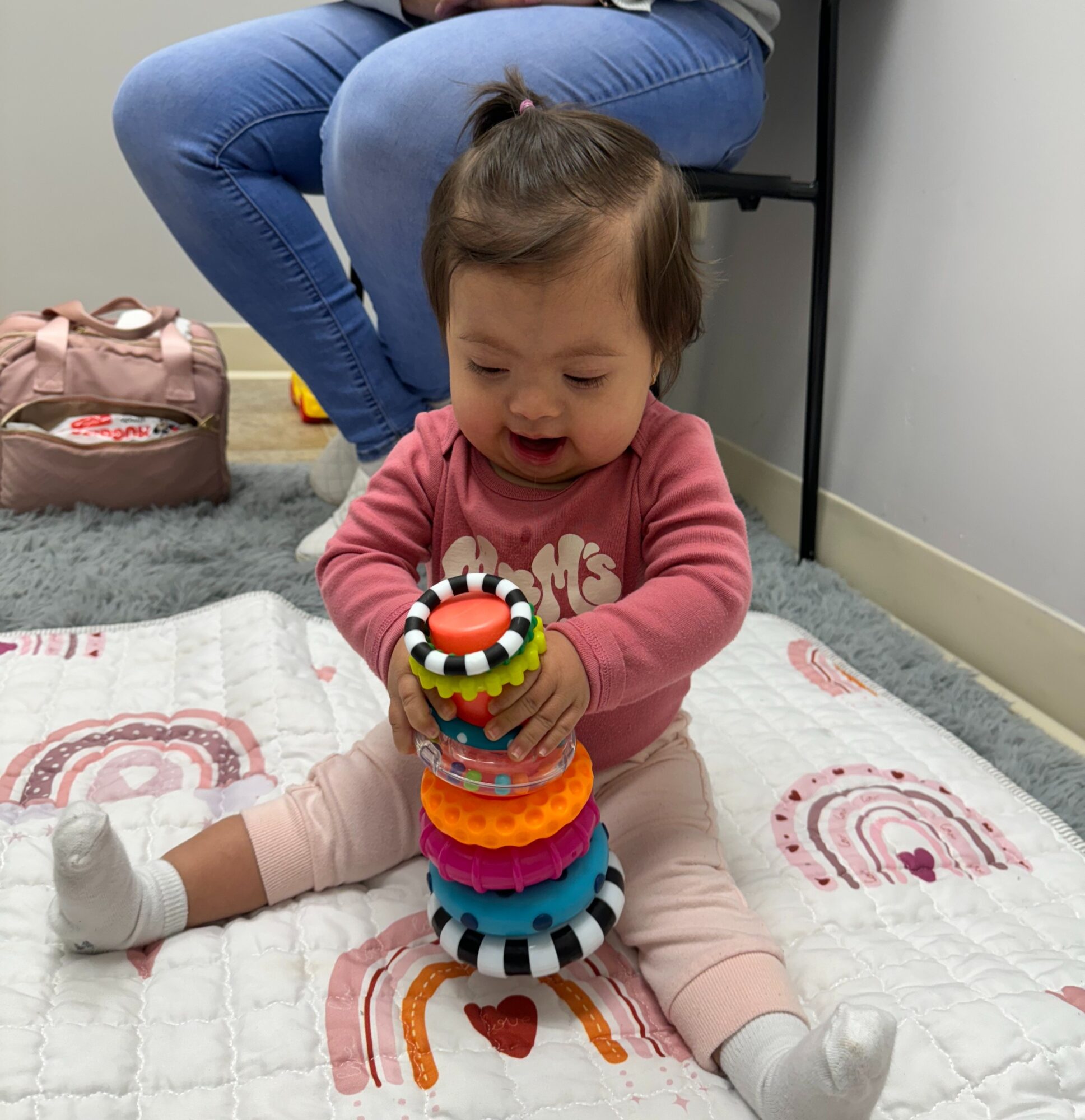 Young girl sitting on a quilted mat, playing with colorful stacking rings, with an adult sitting nearby. Indoor setting.
