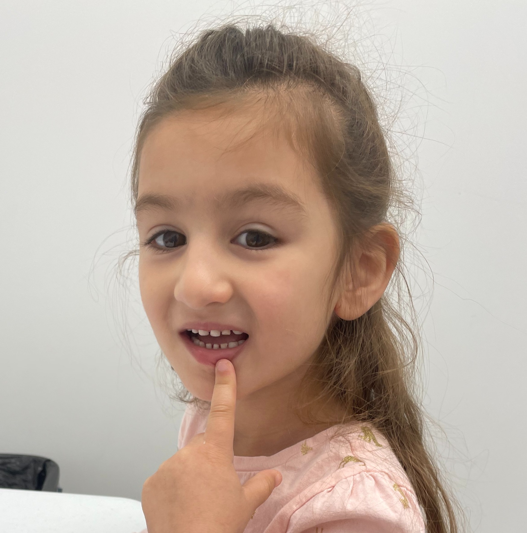 Young girl with light brown hair tied back, touching her lips with her finger, smiling slightly, against a plain white background.