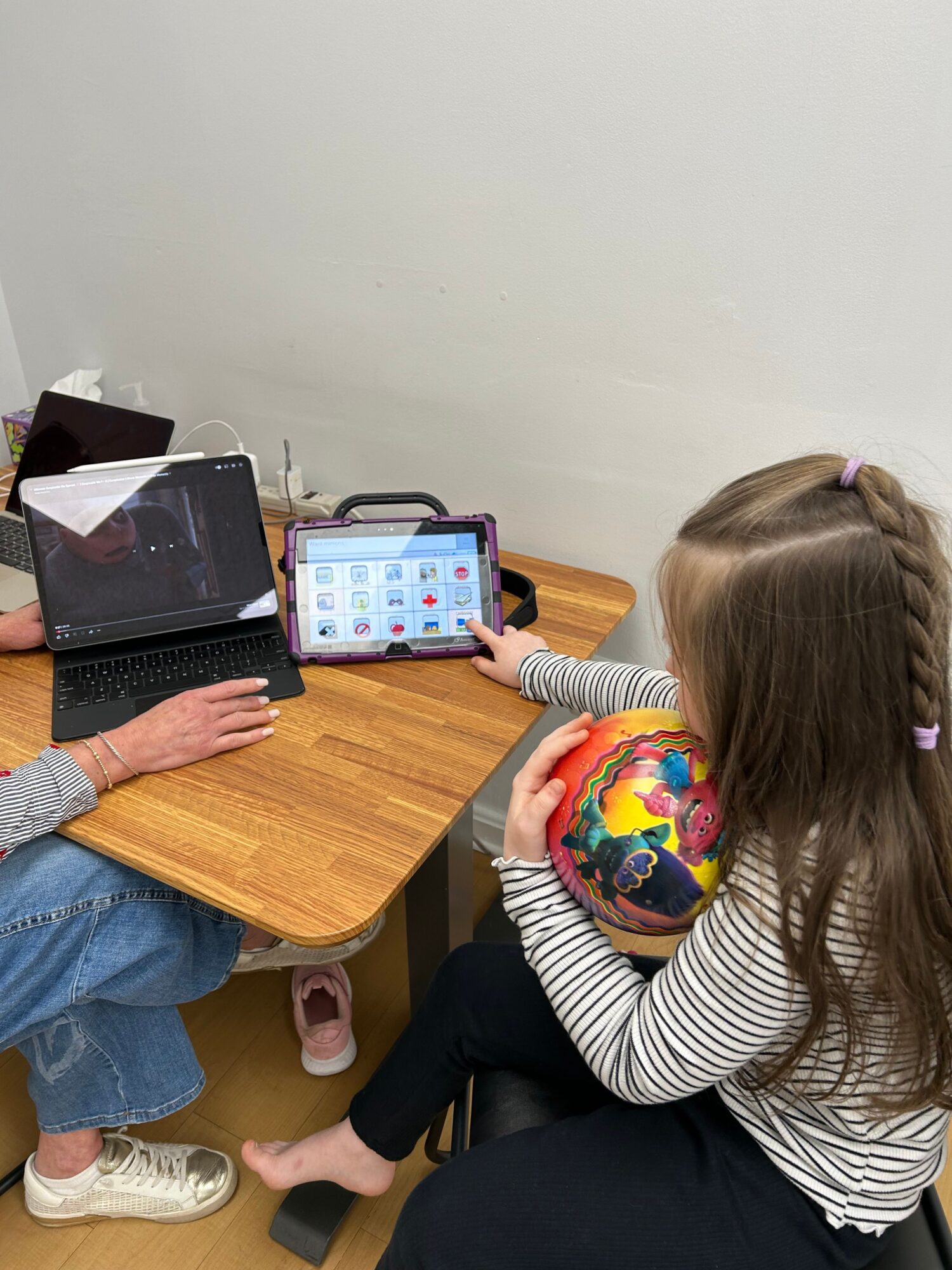 Child with long hair and striped shirt reaches for a tablet on a wooden table, with an adult's hand nearby.