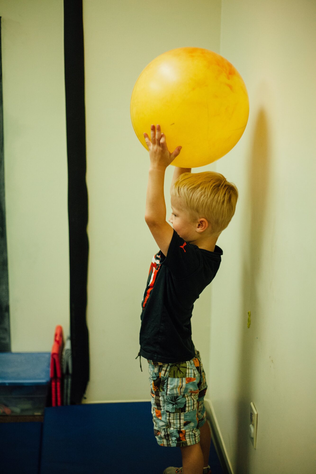 Young boy with blond hair holding a yellow ball above his head, standing indoors near a wall.