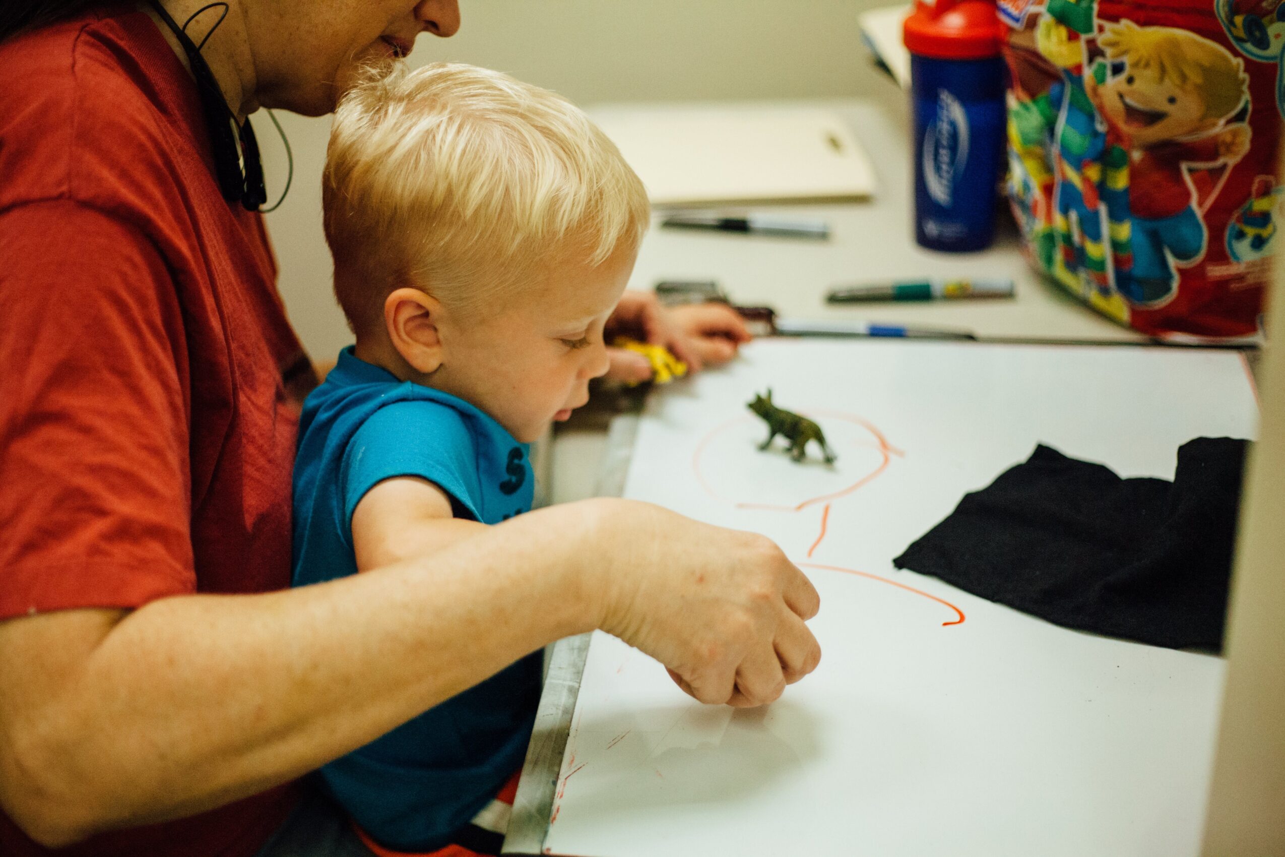 Child and adult drawing on paper with markers, colorful bag and supplies on table, child focused on drawing.