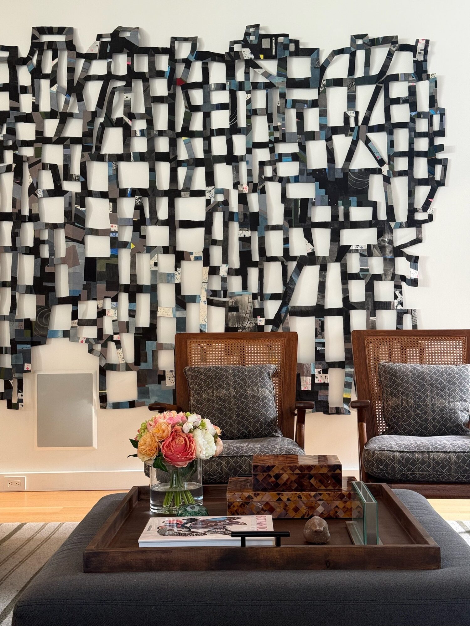 Interior with a wall art installation, a table with a flower arrangement, and two chairs, in a modern setting.