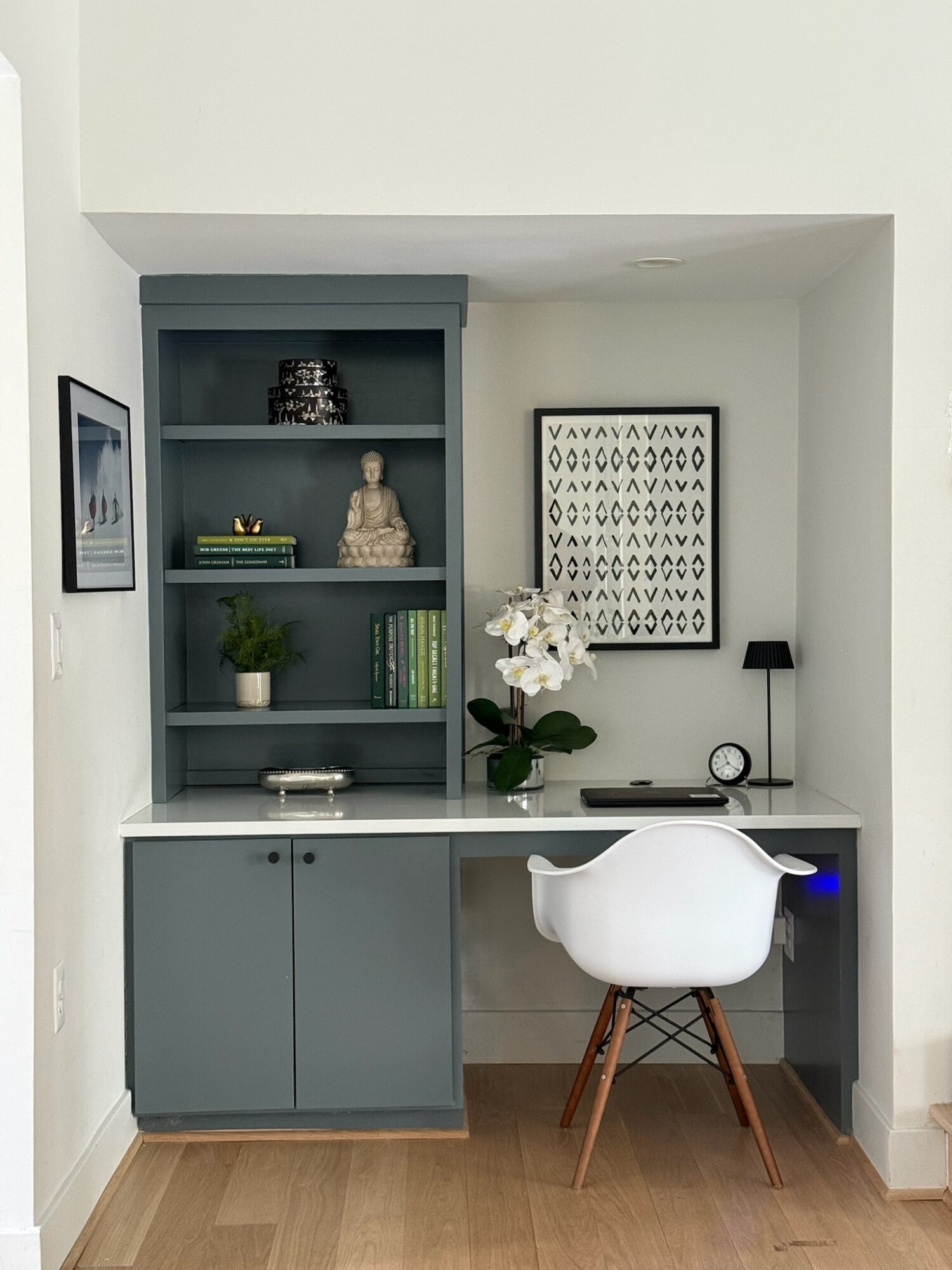 Home office with built-in desk, shelves, white chair, and decorative items, including a Buddha statue and framed artwork, against white walls.