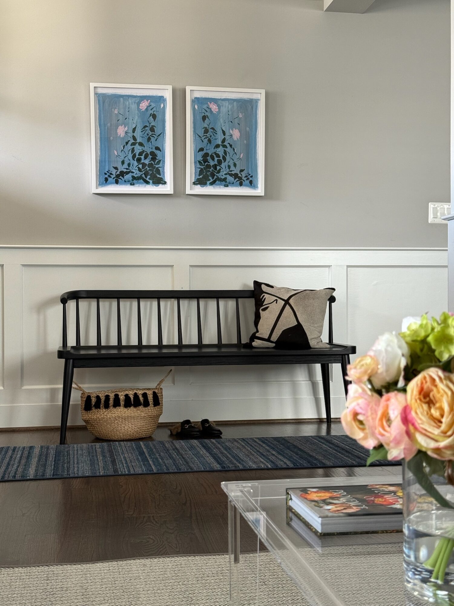 Black bench with a pillow, basket, and shoes in front of a white wall with two framed blue floral artworks, and a glass table with flowers and books.