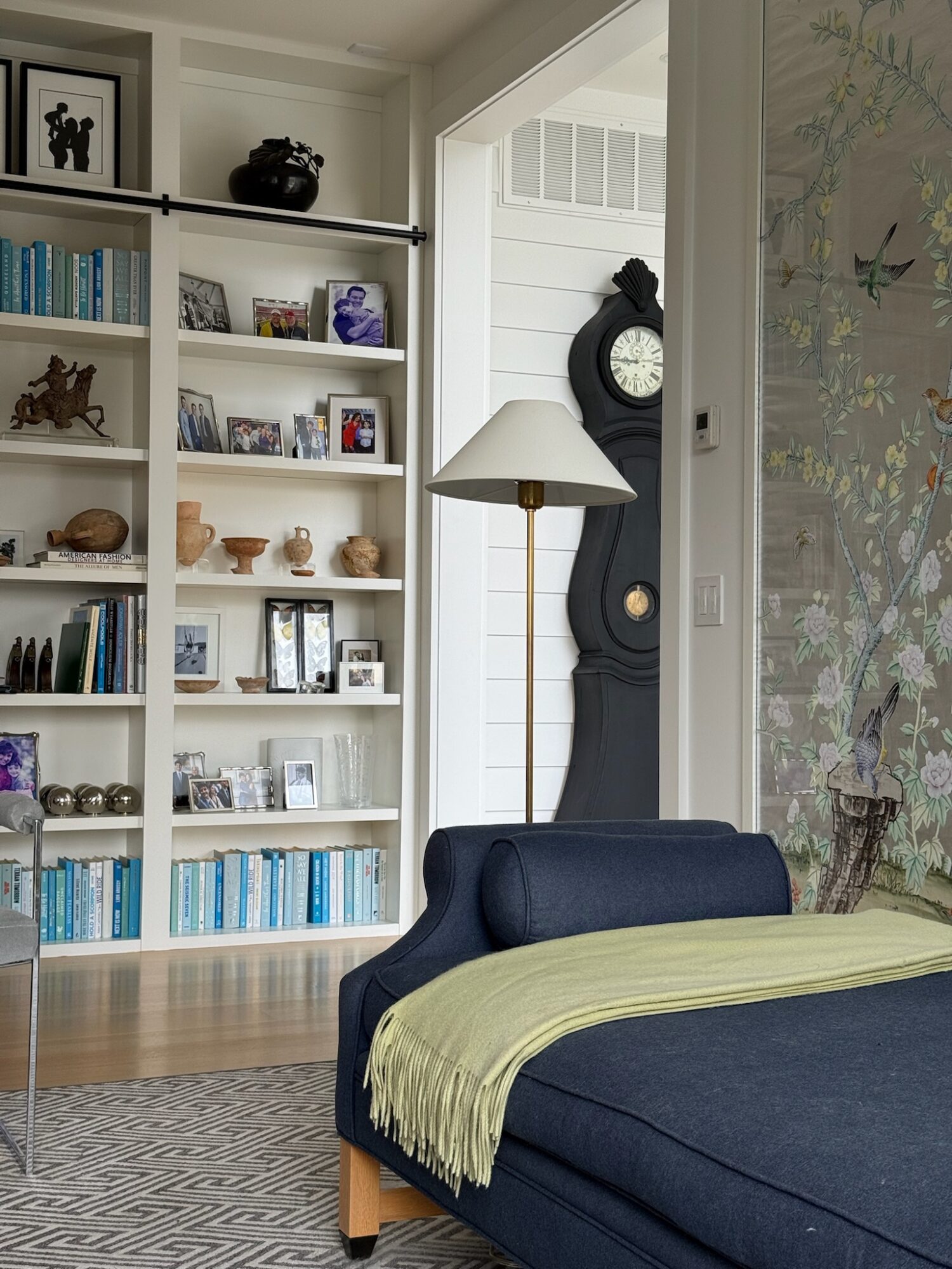 Living room with a blue sofa, white bookshelf, and a tall black clock near a window, with a patterned rug on the floor.