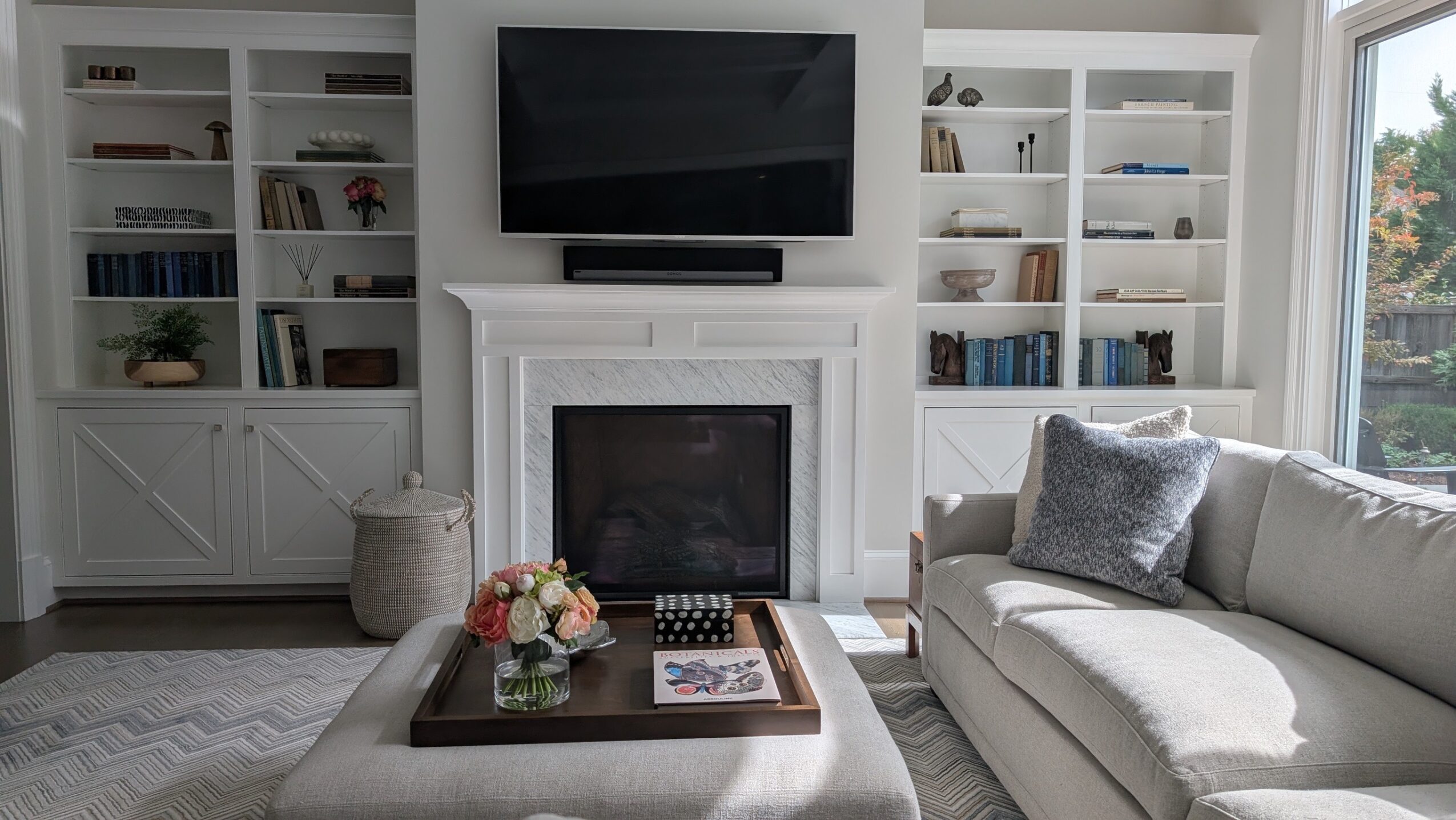 Living room with fireplace, TV, bookshelves, sofa, and window, decorated in neutral tones with cushions and a tray on the ottoman.