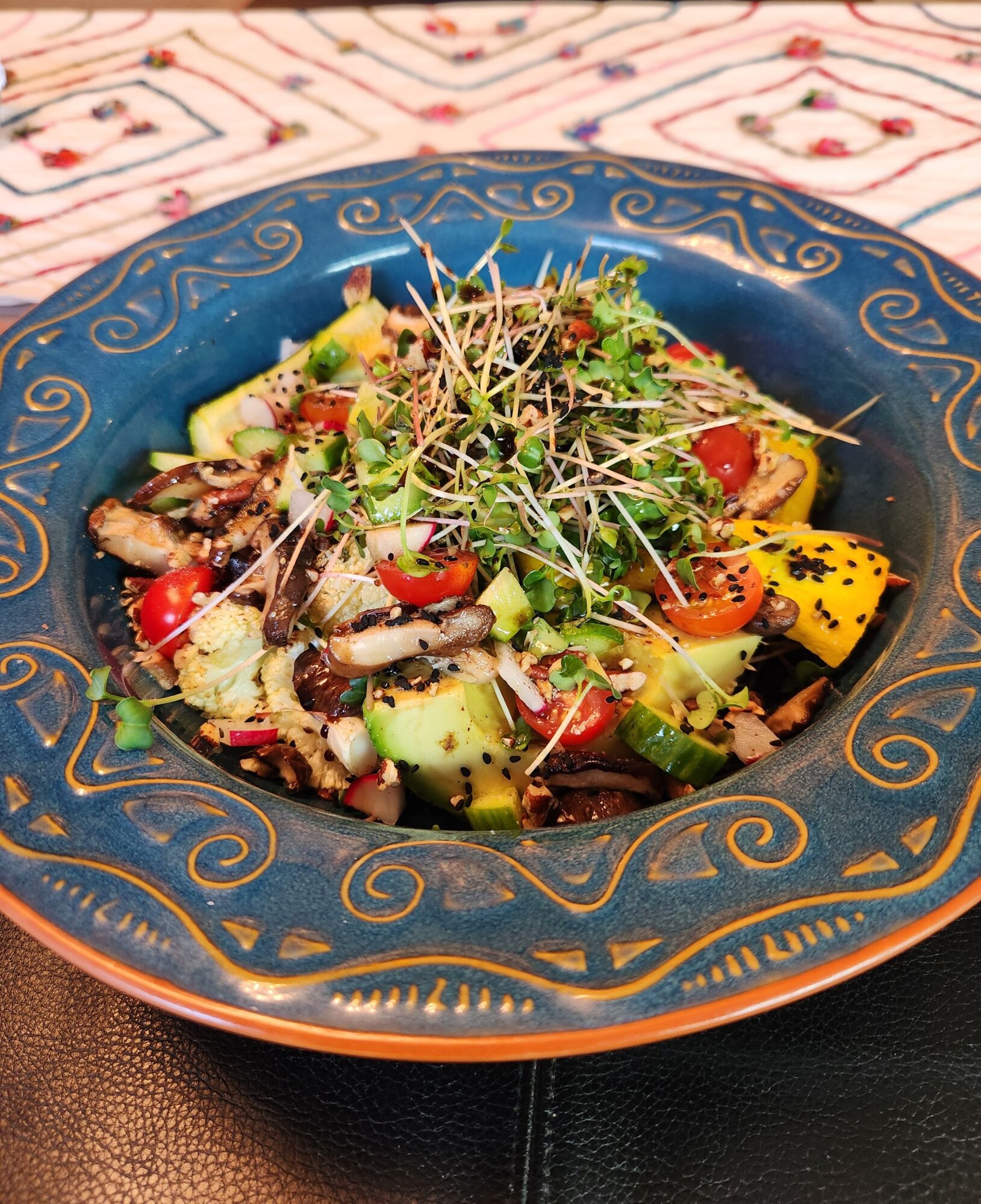 Colorful salad with vegetables and microgreens in a decorative blue bowl on a patterned tablecloth.