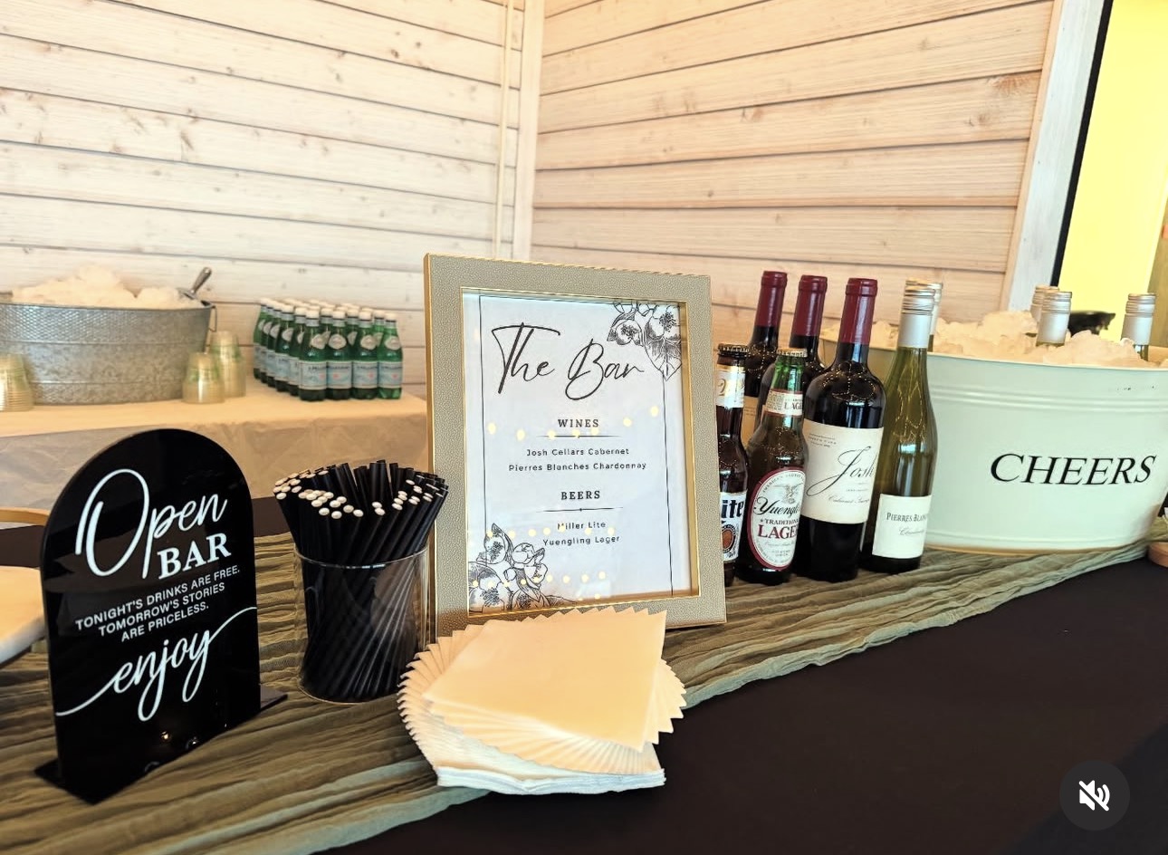 Bar setup with bottles, a sign, napkins, and containers on a wooden counter inside a light wood-paneled room.