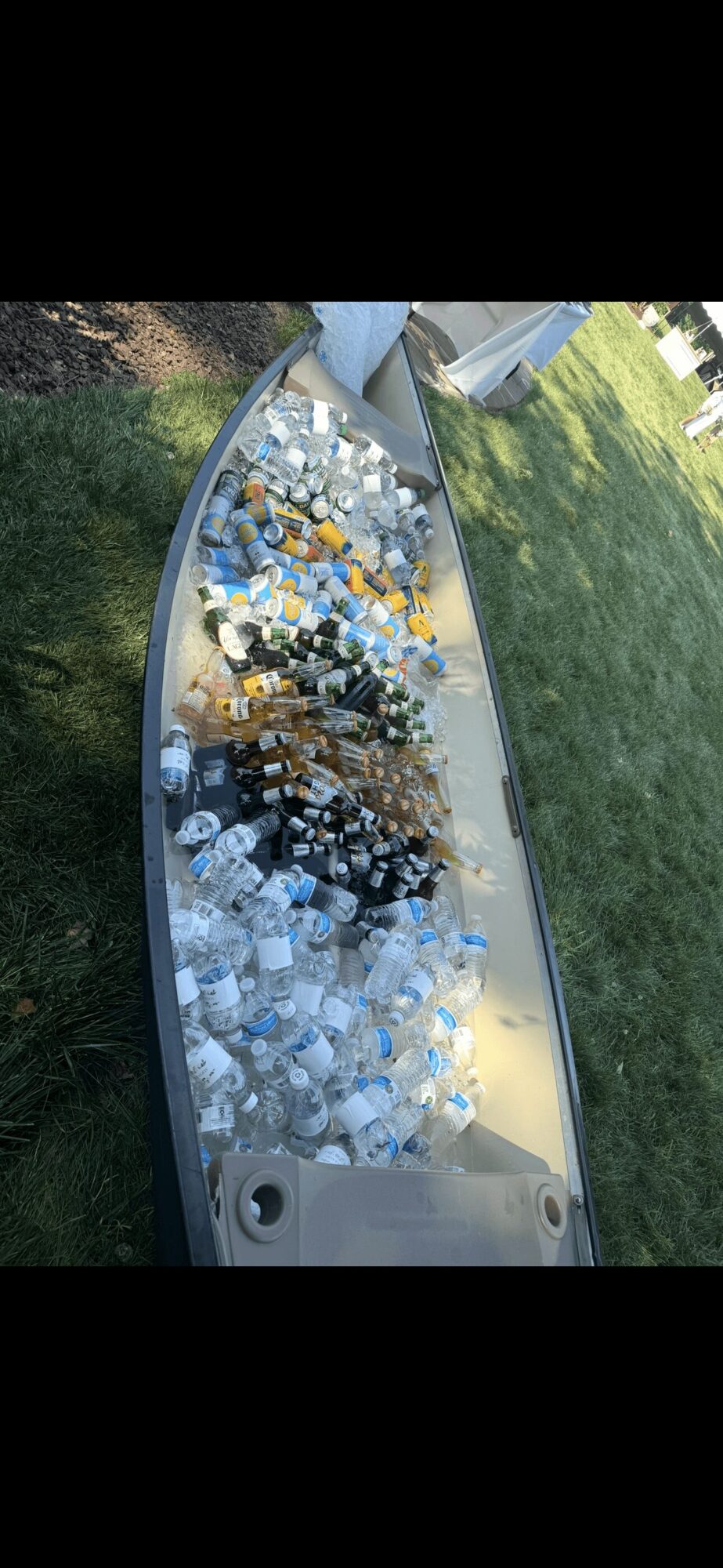 A boat filled with various bottles and containers, on a grassy area with trees and a building in the background.