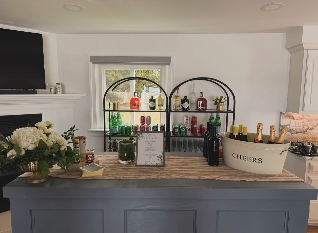 Bar area with shelves holding colorful bottles, a window, and a countertop with flowers and a cheers bucket.