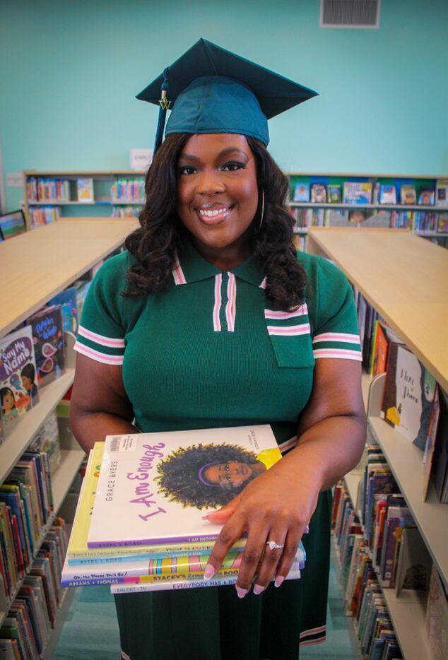 Smiling woman in graduation cap and gown holding a children's book in a library, bookshelves in background.