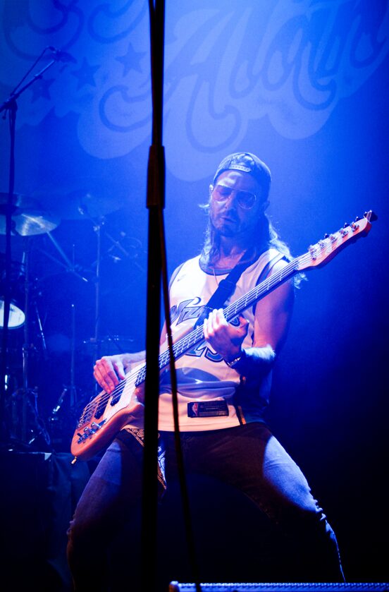 Musician playing an electric guitar on stage with blue lighting and a backdrop, focused and energetic performance.