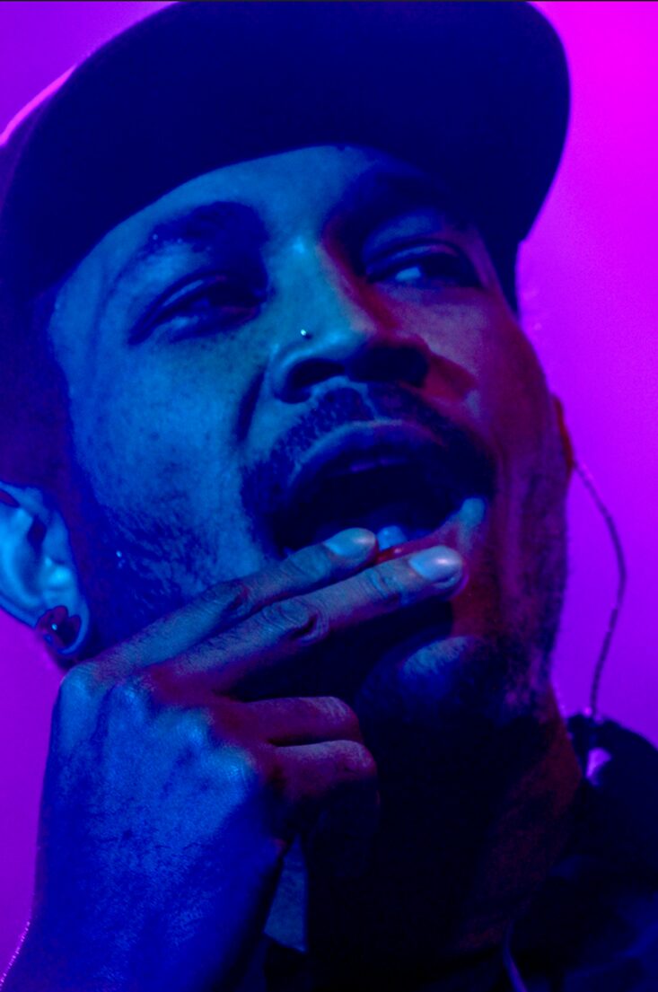 Person with a hat, touching their face, in colorful lighting, looking at the camera.