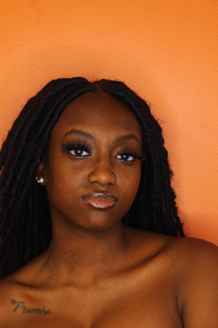 Close-up of a woman with long black dreadlocks, makeup, and a nose piercing, against an orange background.