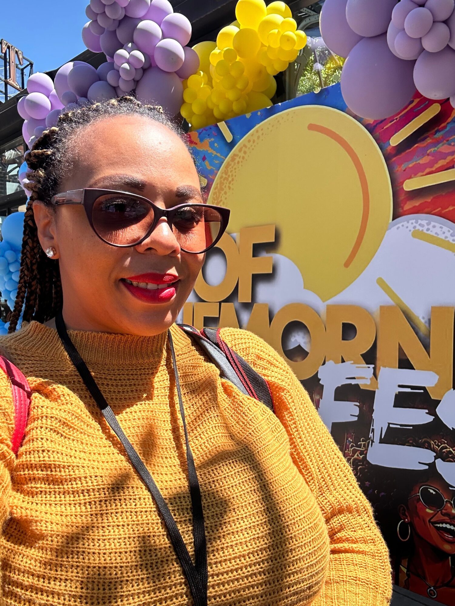 Smiling woman with sunglasses and red lipstick, wearing a yellow sweater, outdoors with colorful balloons and a sun sign in background.