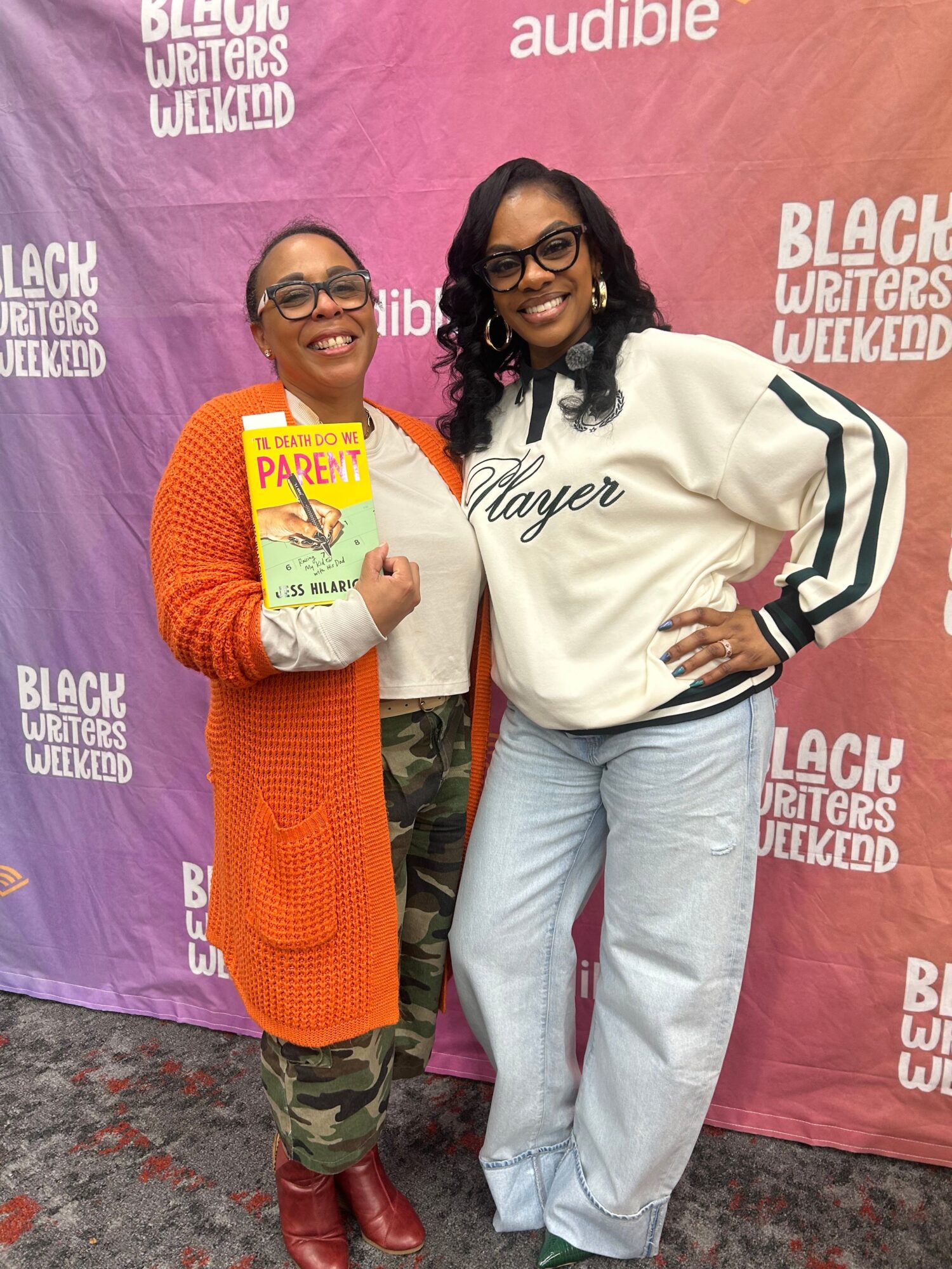 Two women standing in front of a pink and purple backdrop, one holding a book, both smiling, casual clothing, one with glasses.