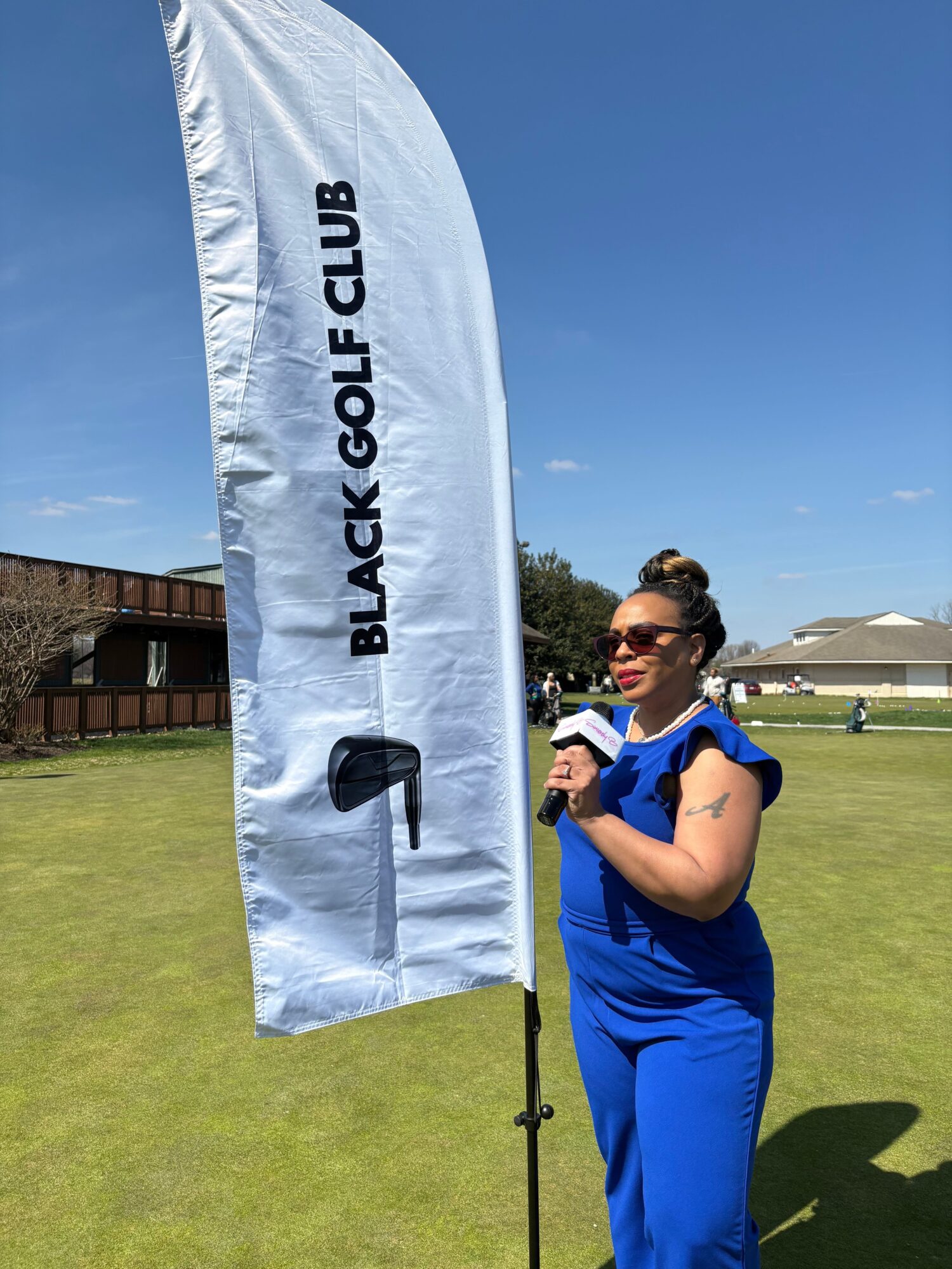 A woman in blue scrubs holding a phone stands on a golf course near a white flag with black text, under a blue sky.