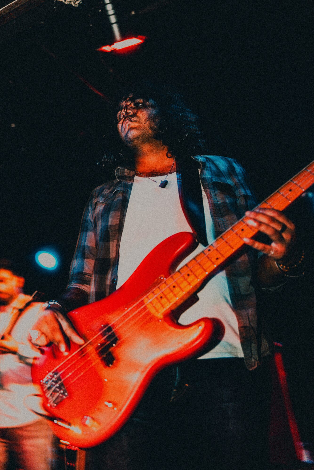 Person playing a red electric bass guitar on stage, wearing glasses and a plaid shirt, with dark background.