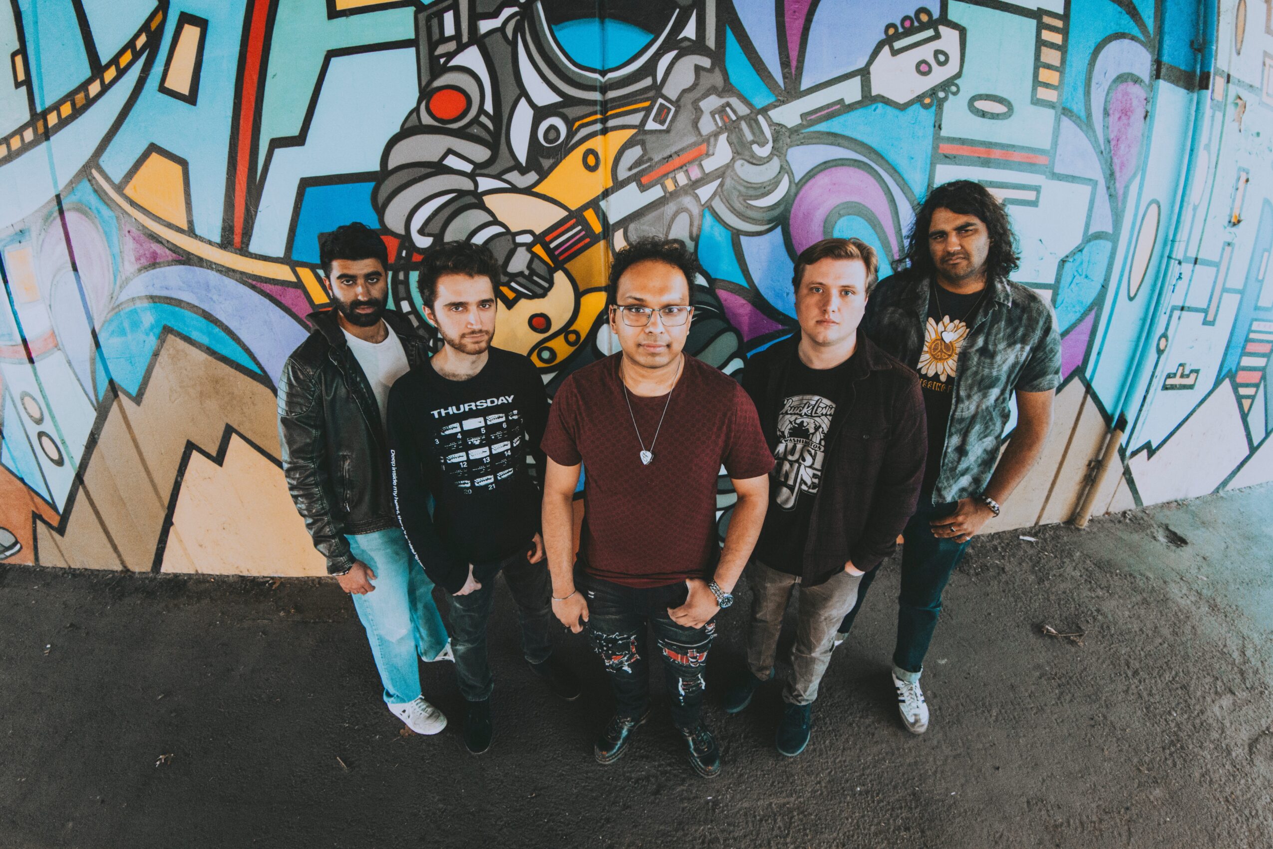 Five people standing in front of colorful graffiti wall, posing for photo, some with hands in pockets, casual clothing.