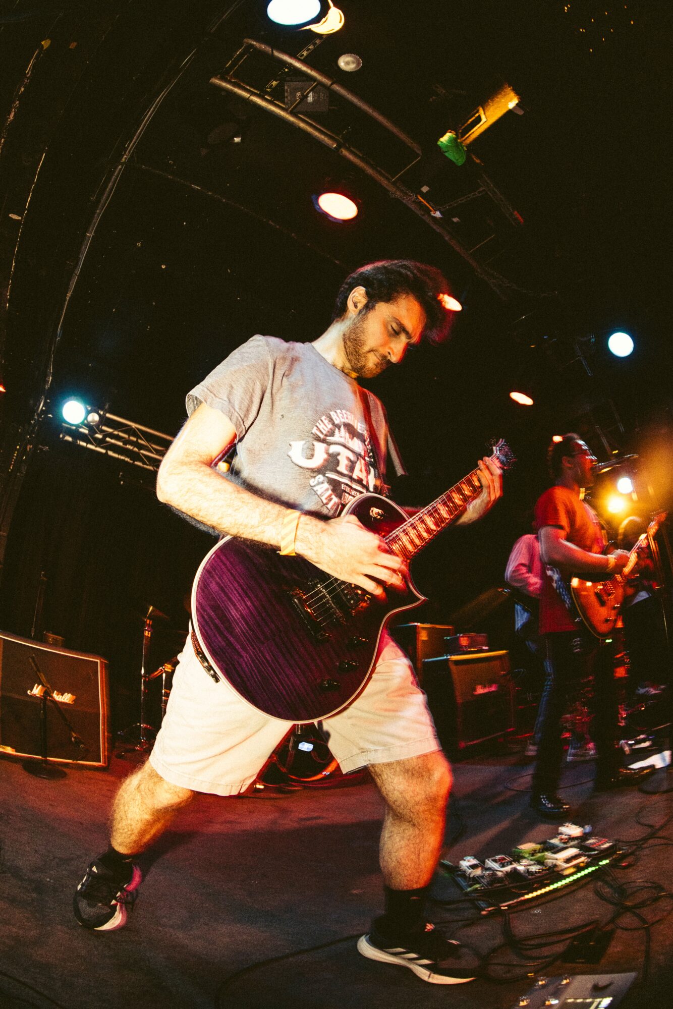 Person playing an electric guitar on stage with stage lights and other band members in the background.