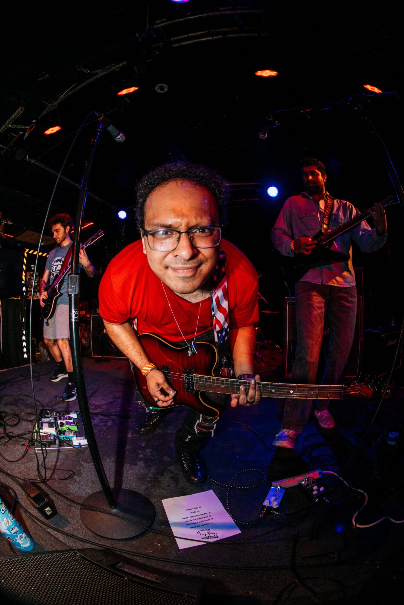 Musician with glasses and red shirt playing guitar on stage with two other band members, stage lights in background.
