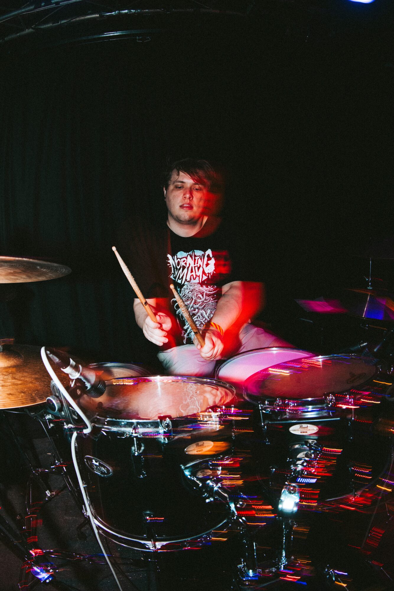 Person playing drums on stage with dark background, holding drumsticks, wearing a black shirt, illuminated by stage lights.