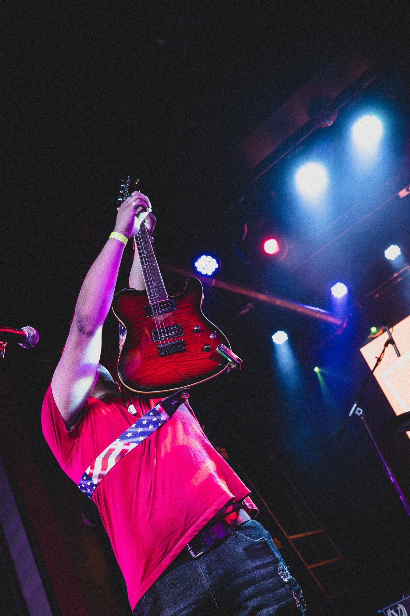 Person holding a red electric guitar on stage with colorful lights, wearing a red shirt, arm raised.