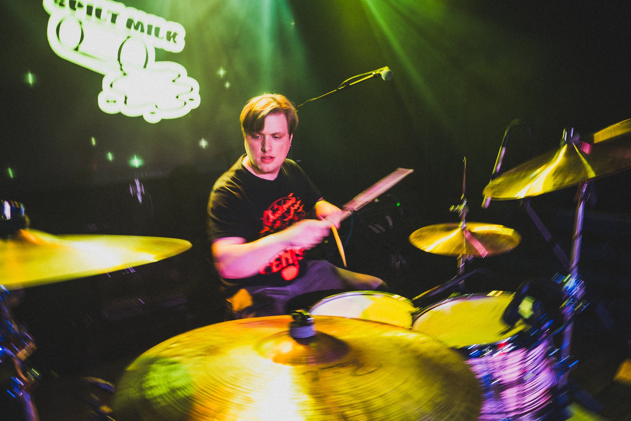 Person playing drums on stage with green and yellow lighting, and a projected logo in the background.