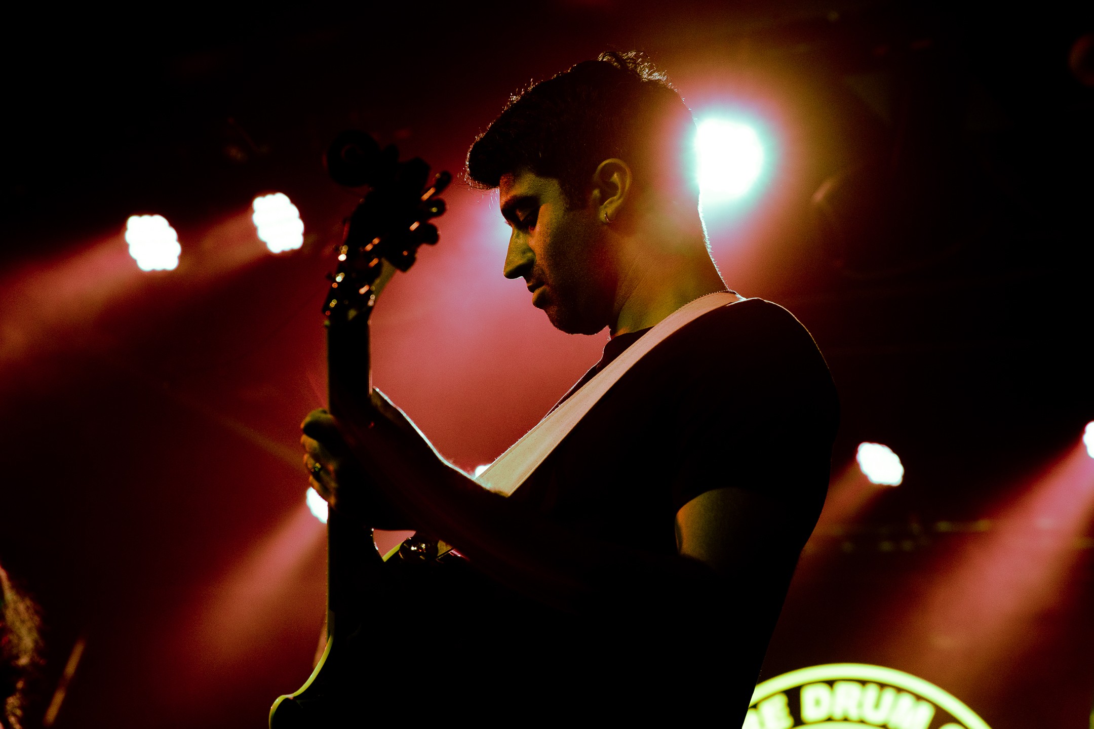 Person playing guitar on stage with bright backlight, dark background, and stage lights. 20 words.