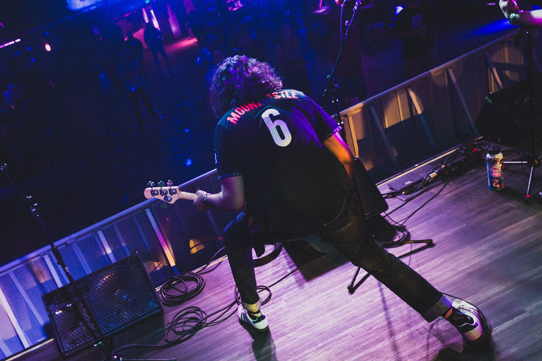Person playing guitar on stage with blue and purple lighting, wearing a black shirt with the number 6, and black pants.